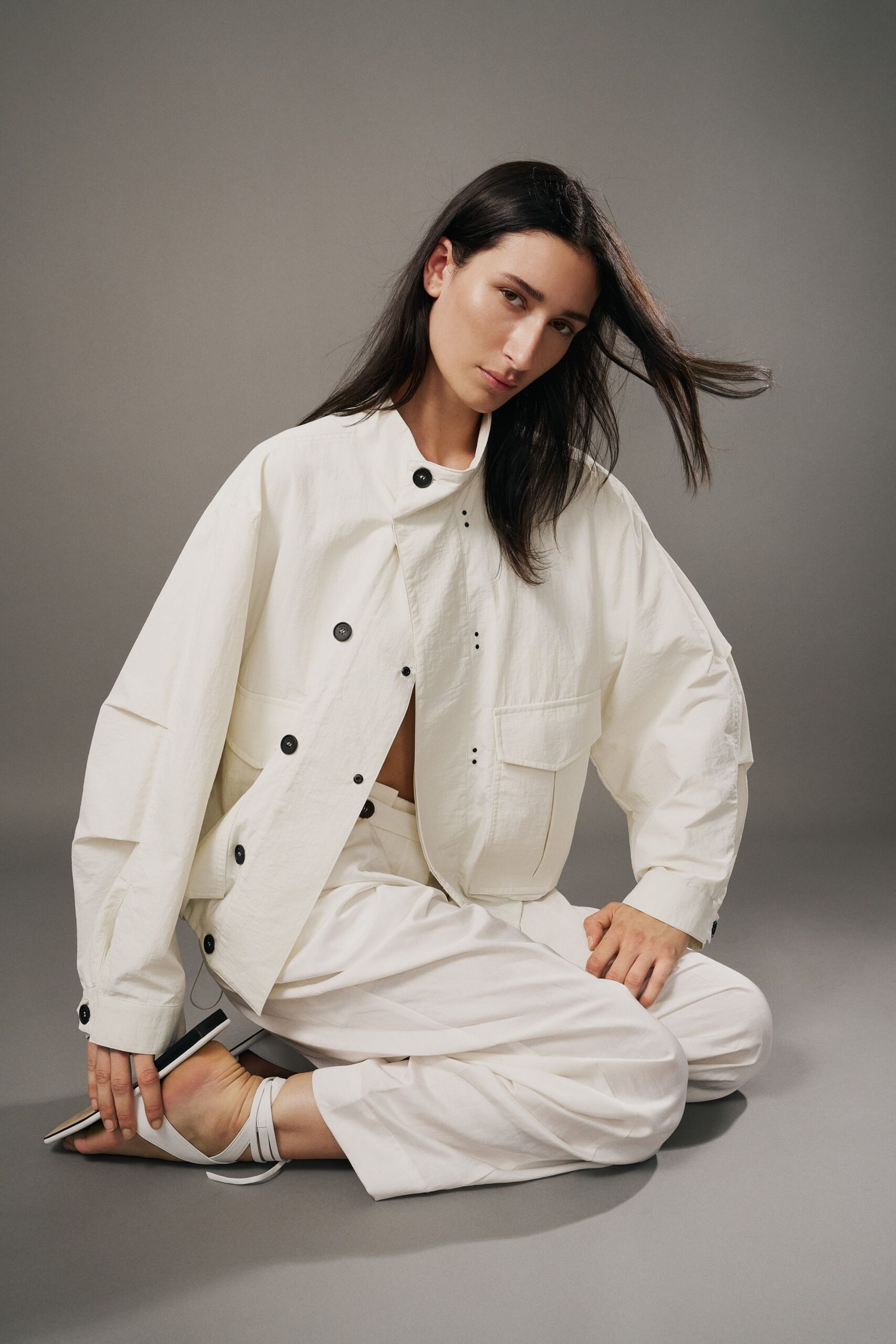 Chalk crinkled jacket with offset black buttons and tiny punched eyelets. Loose chalk trousers with a high tabbed waist and soft front pleats. White wrap sandals, thin straps crossing low at the ankle. Slim black-and-white clutch tucked under the hand. Waiting through a ferry delay with sea mist on the windows.