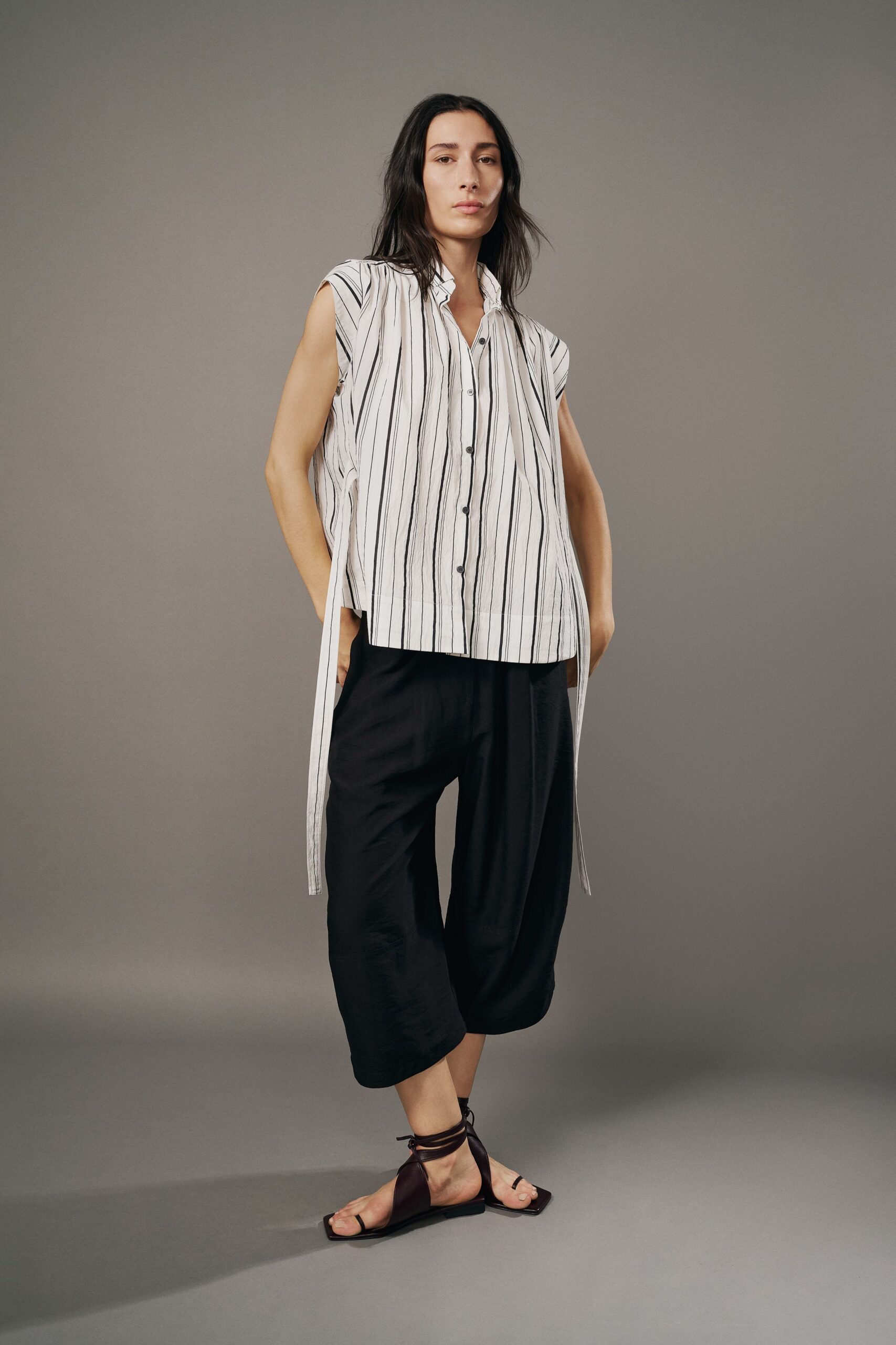White striped cotton blouse with a ruffled collar and long side ties. Black cropped culottes in a fluid weave, ballooning softly at the calf. Dark leather wrap sandals with a squared sole and toe loop. Walking across a stone courtyard before the afternoon heat settles in.