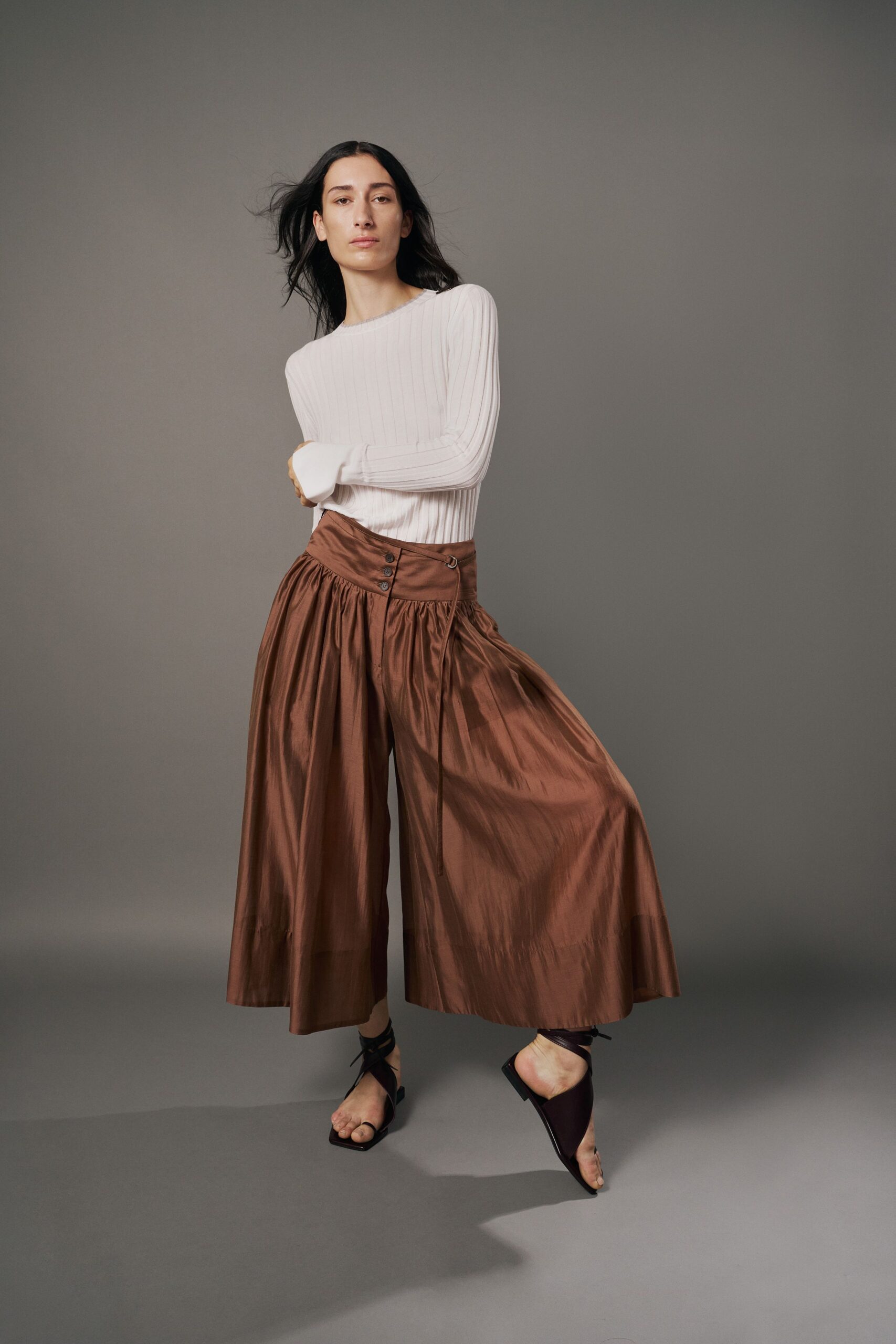 Ivory ribbed knit top with a narrow crew neck. Coppery taffeta culottes, softly gathered under a slanted waistband with four buttons. Dark wrap sandals laced high at the ankle. Walking across a museum atrium before the doors open.