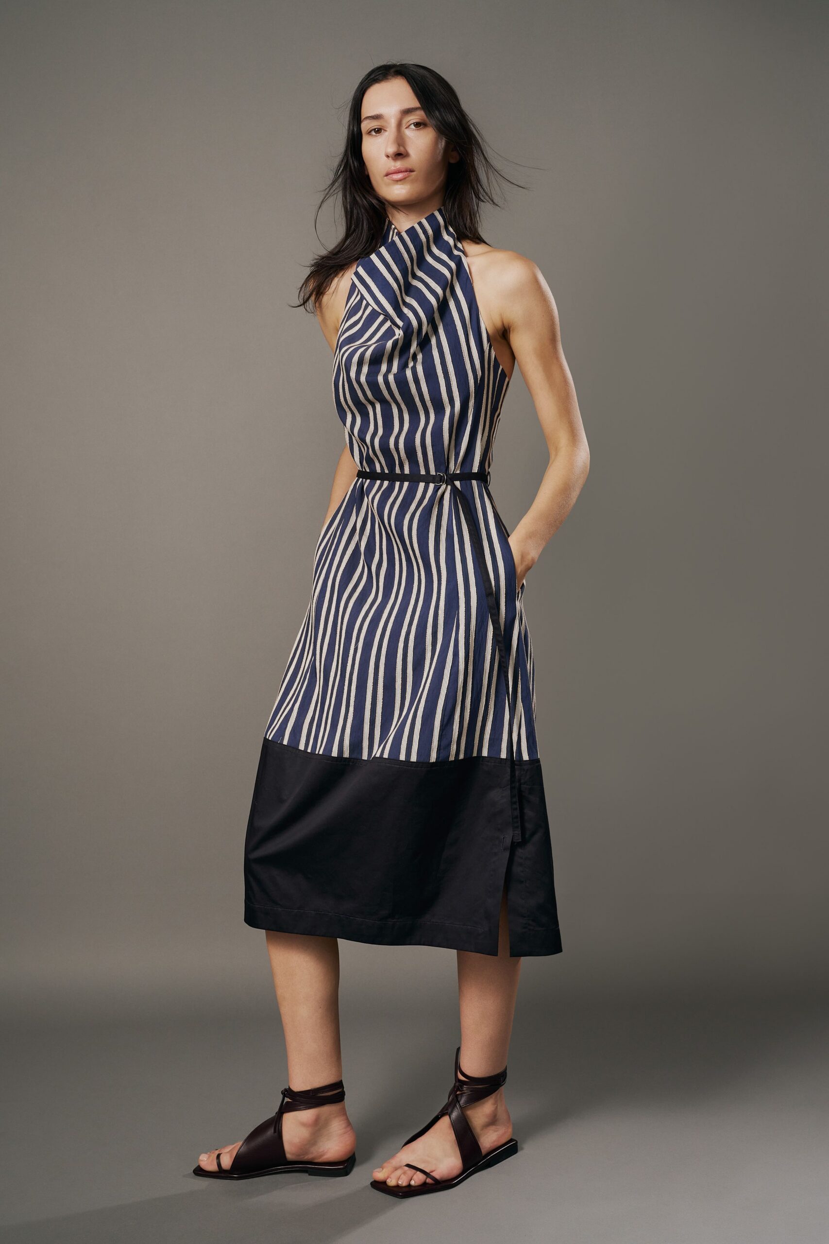 Navy and chalk striped halter dress, neck drape folding into a narrow tie belt. Dark leather wrap sandals, cords crossing twice at the ankle. Walking back from the marina with salt drying on your shoulders.