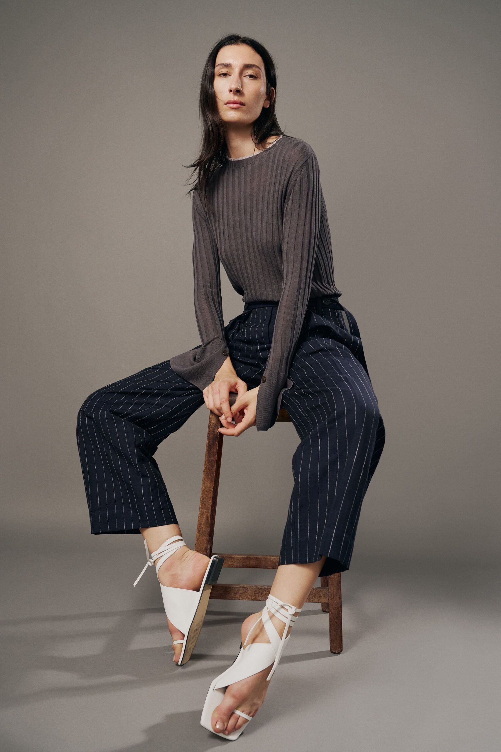 Taupe ribbed knit top with elongated cuffs split at the wrist. Cropped navy trousers in chalk pinstripes, pleated softly at the waist. White square-toe sandals laced high around the ankles. Waiting out a long lunch where the chairs are all too low.