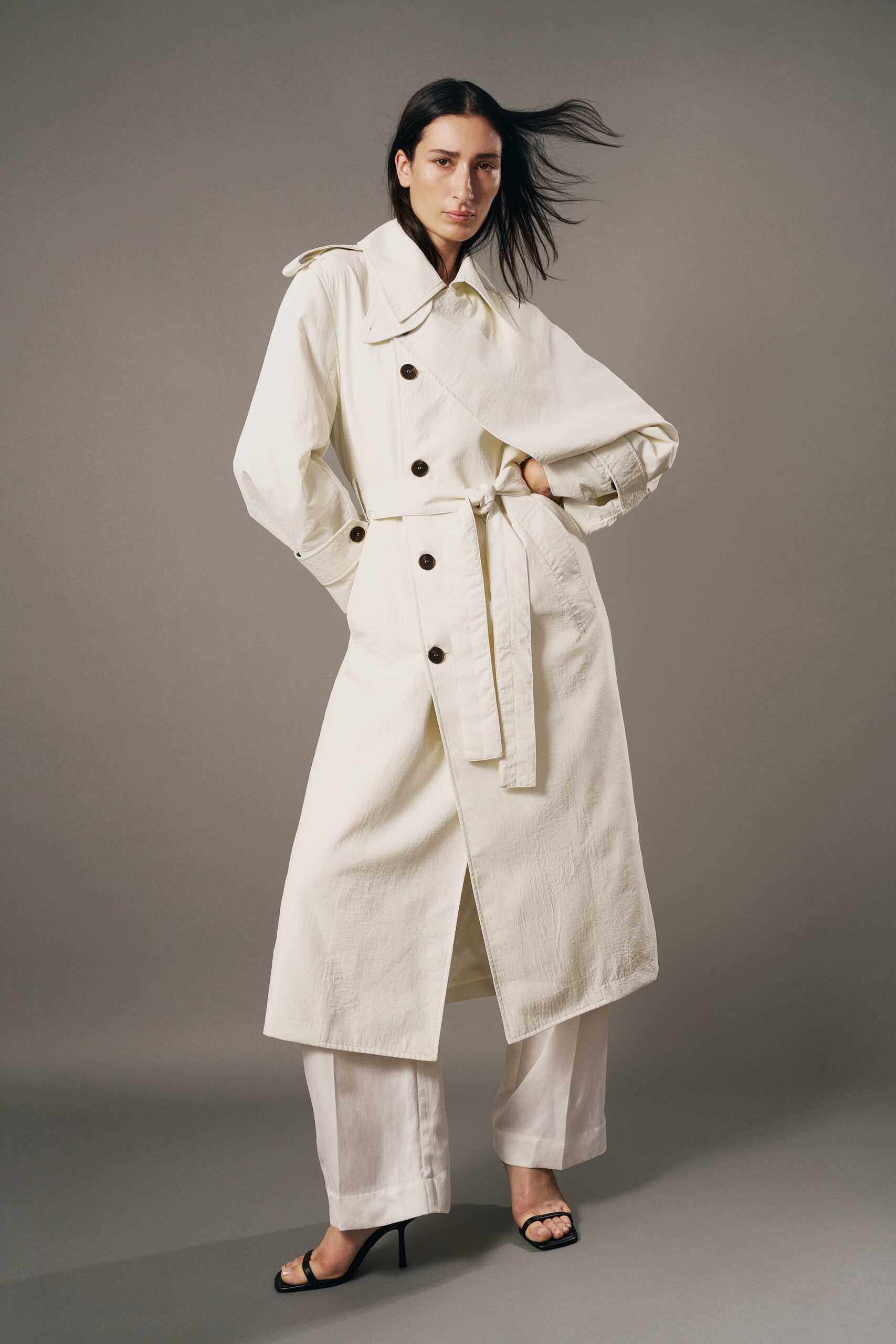 Chalk trench in crisp washed cotton, storm flap skewed over dark buttons. Wide white trousers with deep turned cuffs and a soft break. Black square-toe heeled sandals, bare instep and thin straps. Walking into a windy ferry terminal before the first commuter rush.