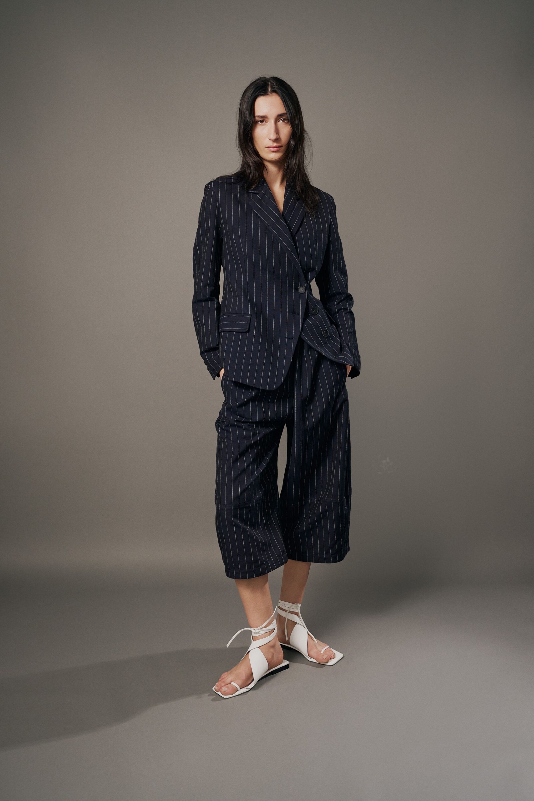 Navy pinstripe blazer with peaked lapels and a slanting double-breasted wrap. Matching pinstripe culottes cut extra wide, breaking in a deep front pleat. White square-toe sandals laced high around the ankles. First meeting in an office still smelling faintly of fresh paint.