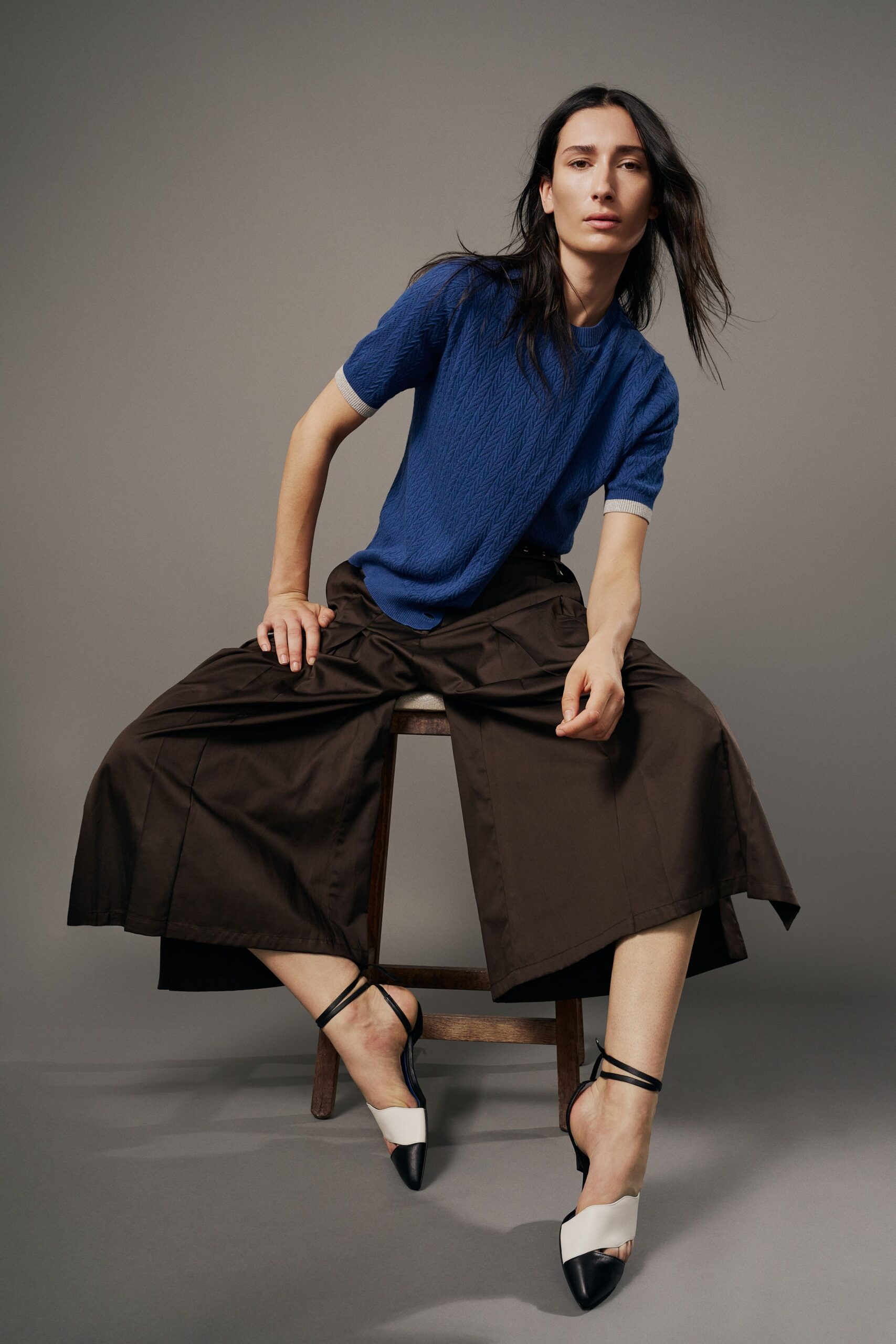Cobalt short-sleeve knit with a narrow chevron texture and pale rib cuffs. Deep brown culottes in crisp cotton, folded side flaps lifting off the hip. Black-and-cream slingbacks with thin ankle straps and pointed cap toes. Waiting through the quiet hour before a museum cafe opens its shutters.