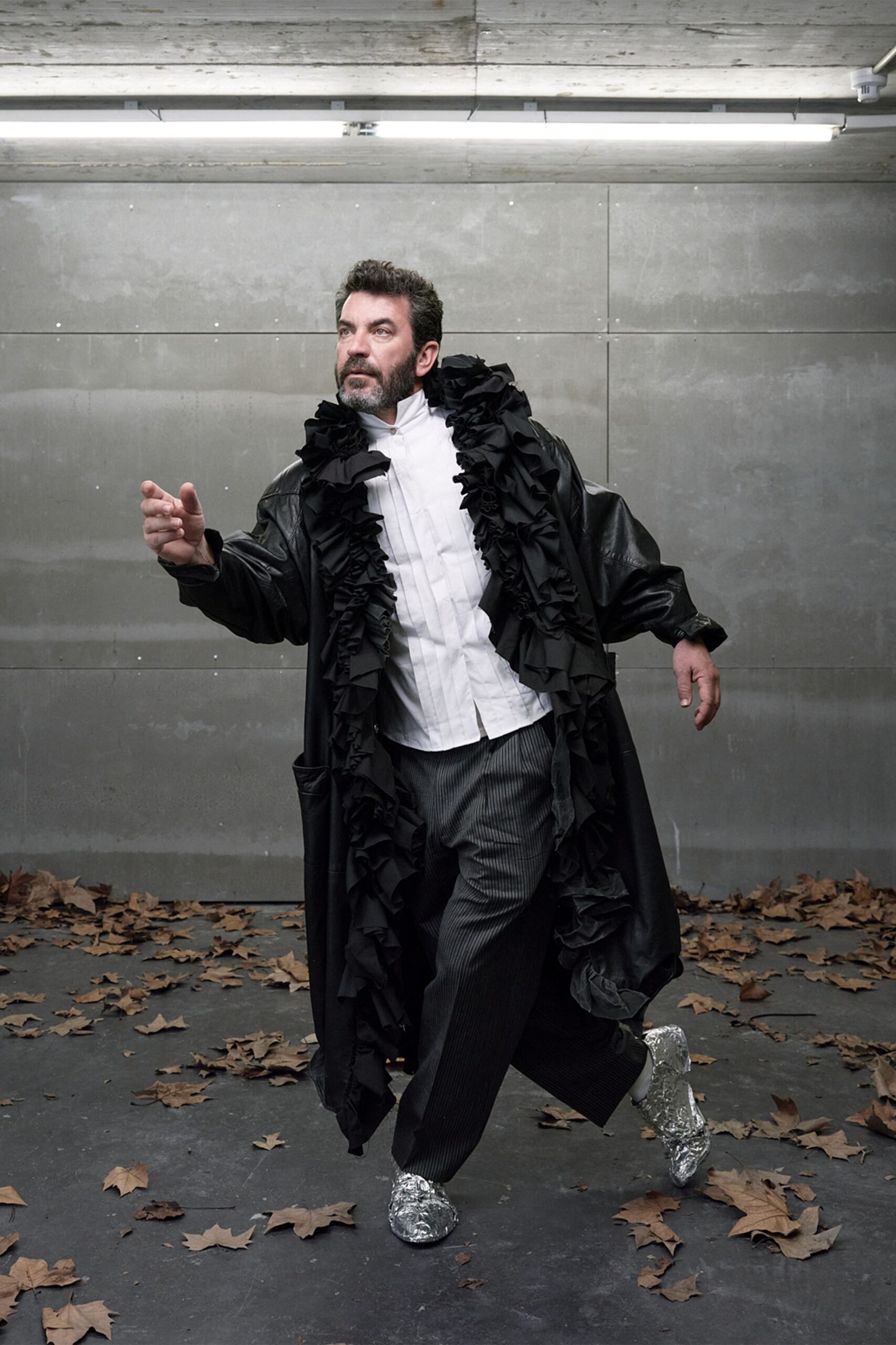 Long black leather coat, front swallowed by piled matte ruffles. White bib-front shirt with narrow pintucks and a stand collar. Charcoal pinstriped trousers, loose through the leg. Silver shoes wrapped in crinkled foil. Crossing a parking garage floor littered with plane-tree leaves after a costume fitting.