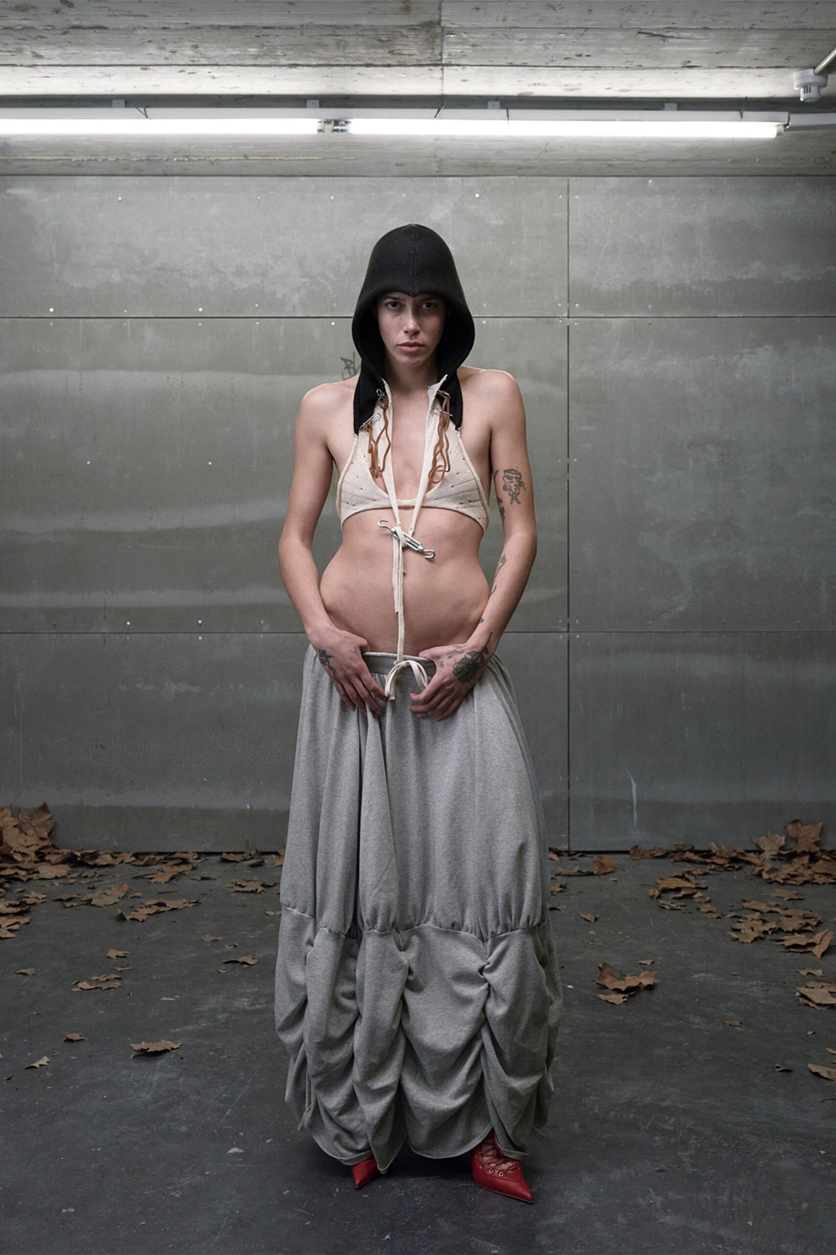 Black felted hood hanging in long earflaps. Bone knit triangle bralette, hooked closed below the ribs. Grey jersey skirt ballooned into gathered swags near the floor. Red pointed mules with raw lacing at the vamp. Under strip lights in a concrete loading bay, leaves blown in after rain.