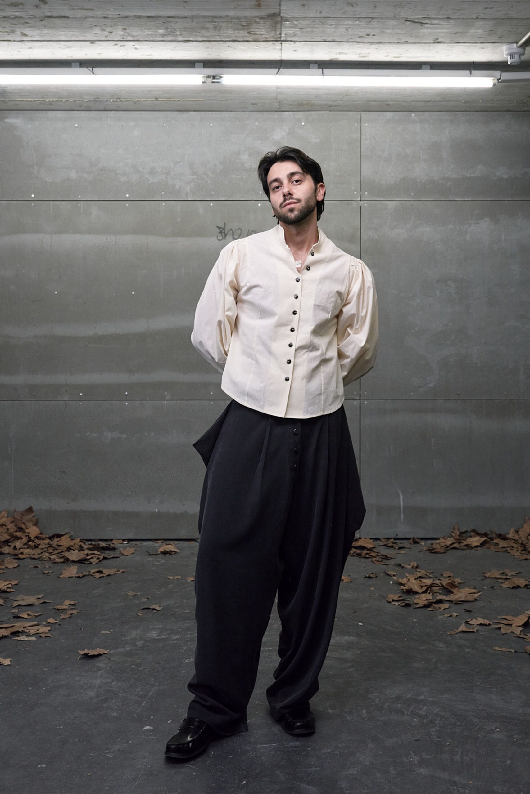 Cream cotton blouse with gathered sleeves and a skewed line of dark buttons. Black voluminous trousers, cut extra wide with a loose panel swinging at one hip. Black leather loafers with squared toes. After-hours in a concrete loading bay, leaves blown into the corners.