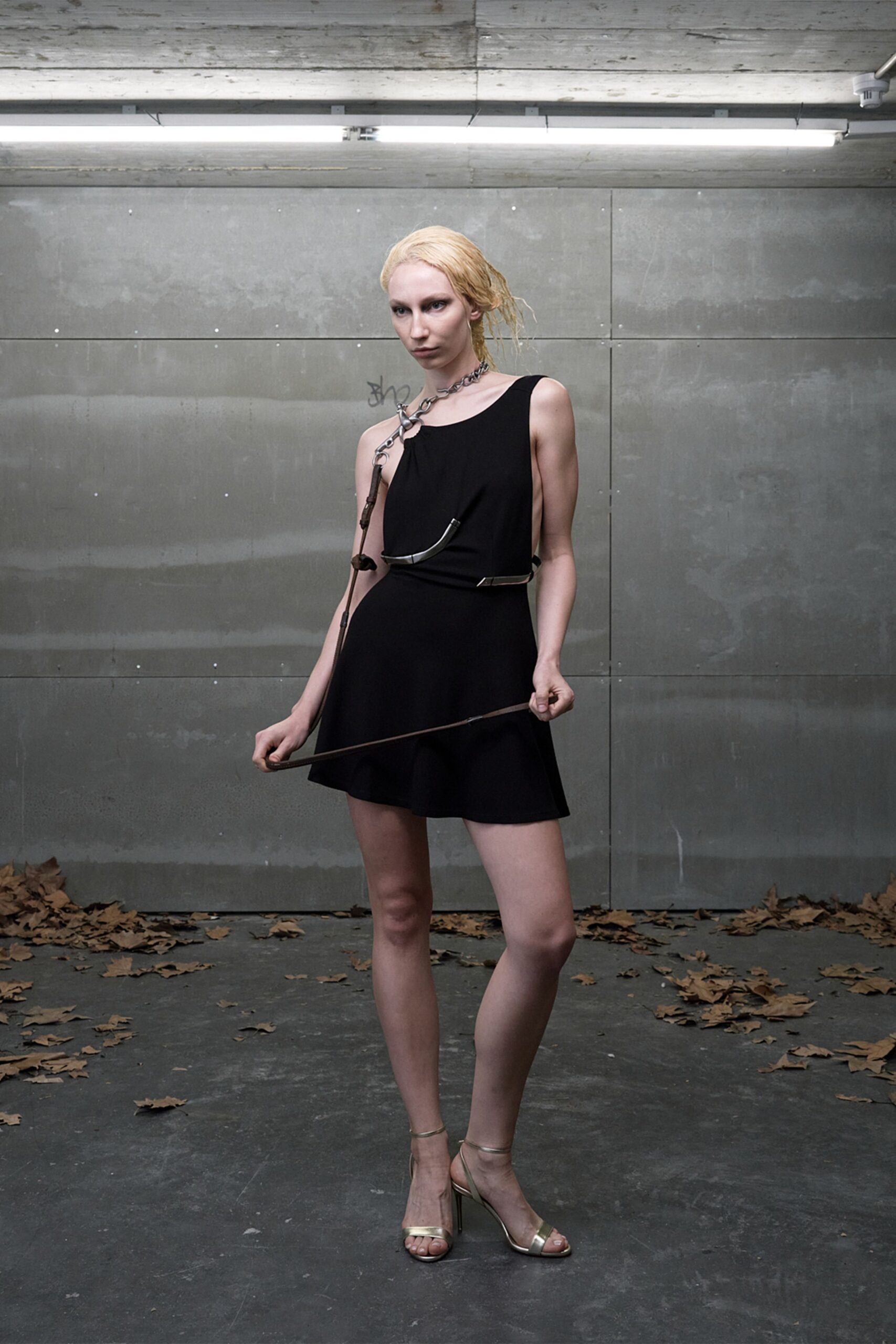 Black mini dress with a low side cut and softly flared hem. Heavy chain collar feeding a brown strap and curved metal tube across the chest. Pale metallic ankle-strap sandals on thin stiletto heels. Under strip lights in a concrete loading bay, leaves blown in after dark.