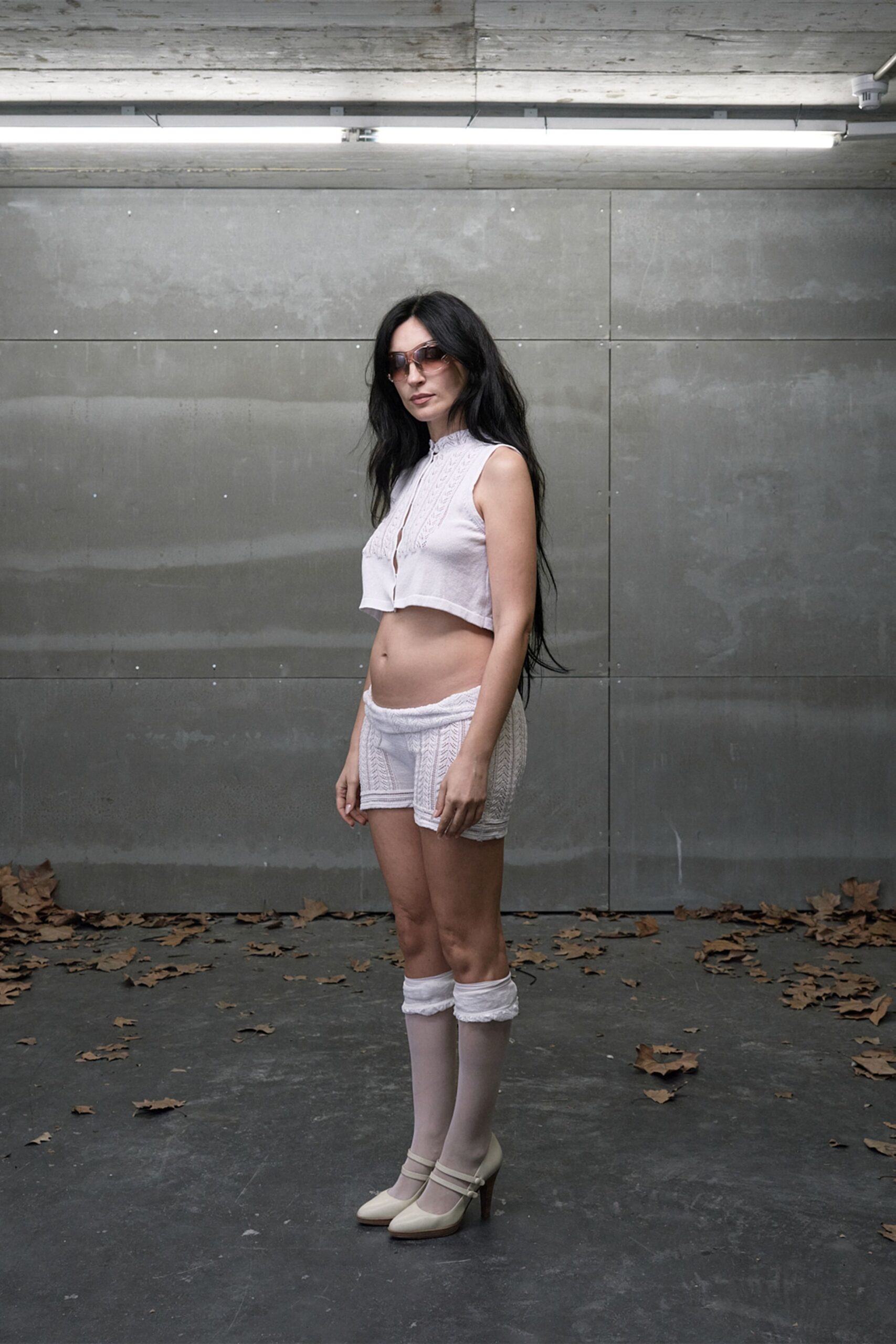 Brown gradient shield sunglasses. White knit cropped top with cable panels and a tiny keyhole slit. Low-slung white knit shorts, rolled waistband exposing the hip line. Sheer white knee socks with ruffled cuffs. Cream double-strap pumps with rounded toes. Under a parking-garage strip light, leaves skittering in from the street.
