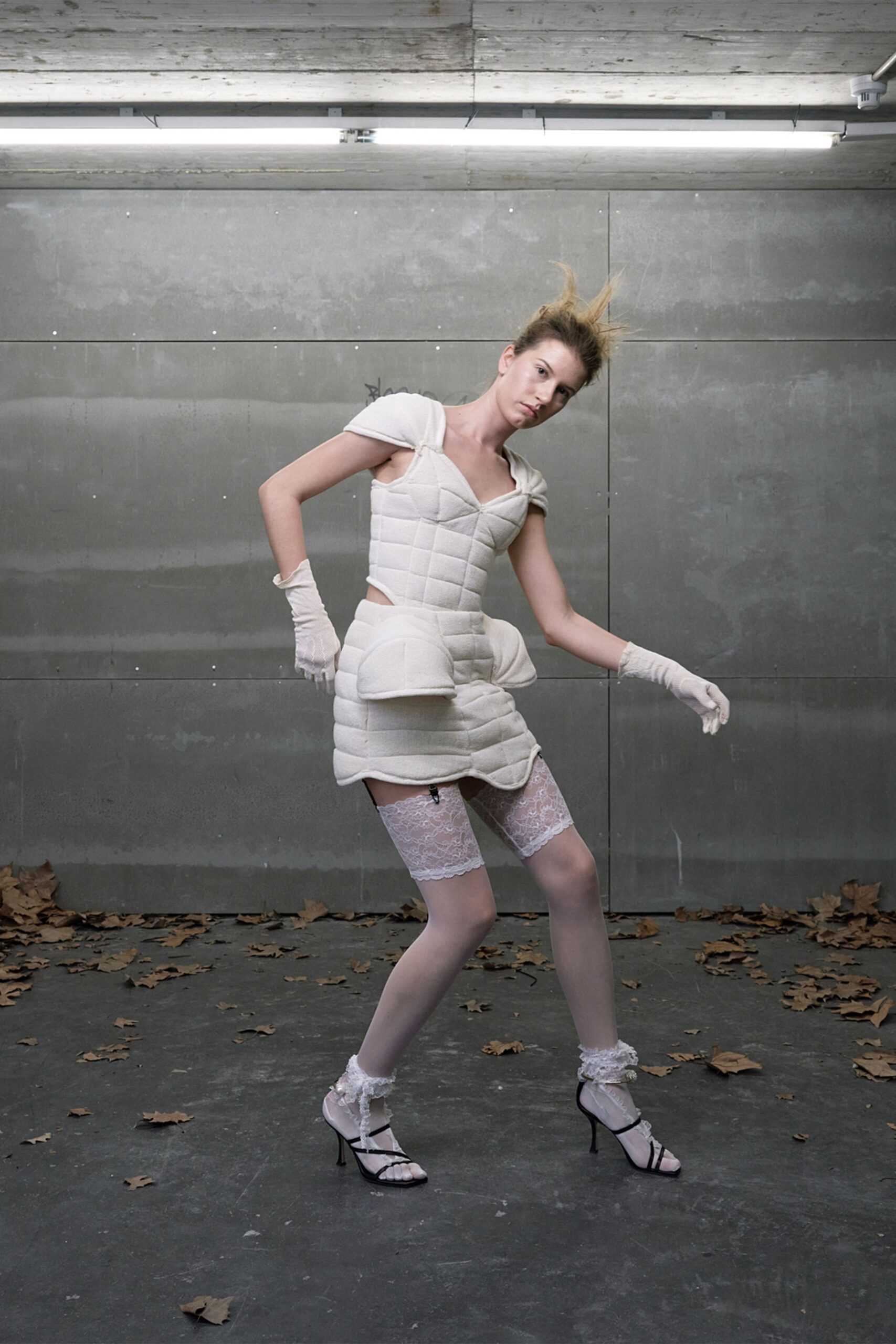 Ivory padded dress, channel-quilted with bulbous hip panniers and a curved hem. Short white mesh gloves. White lace stay-up tops peeking under the skirt. Black strappy heels with frothy lace anklets. Under strip lights in a concrete loading bay, leaves blown in after closing.