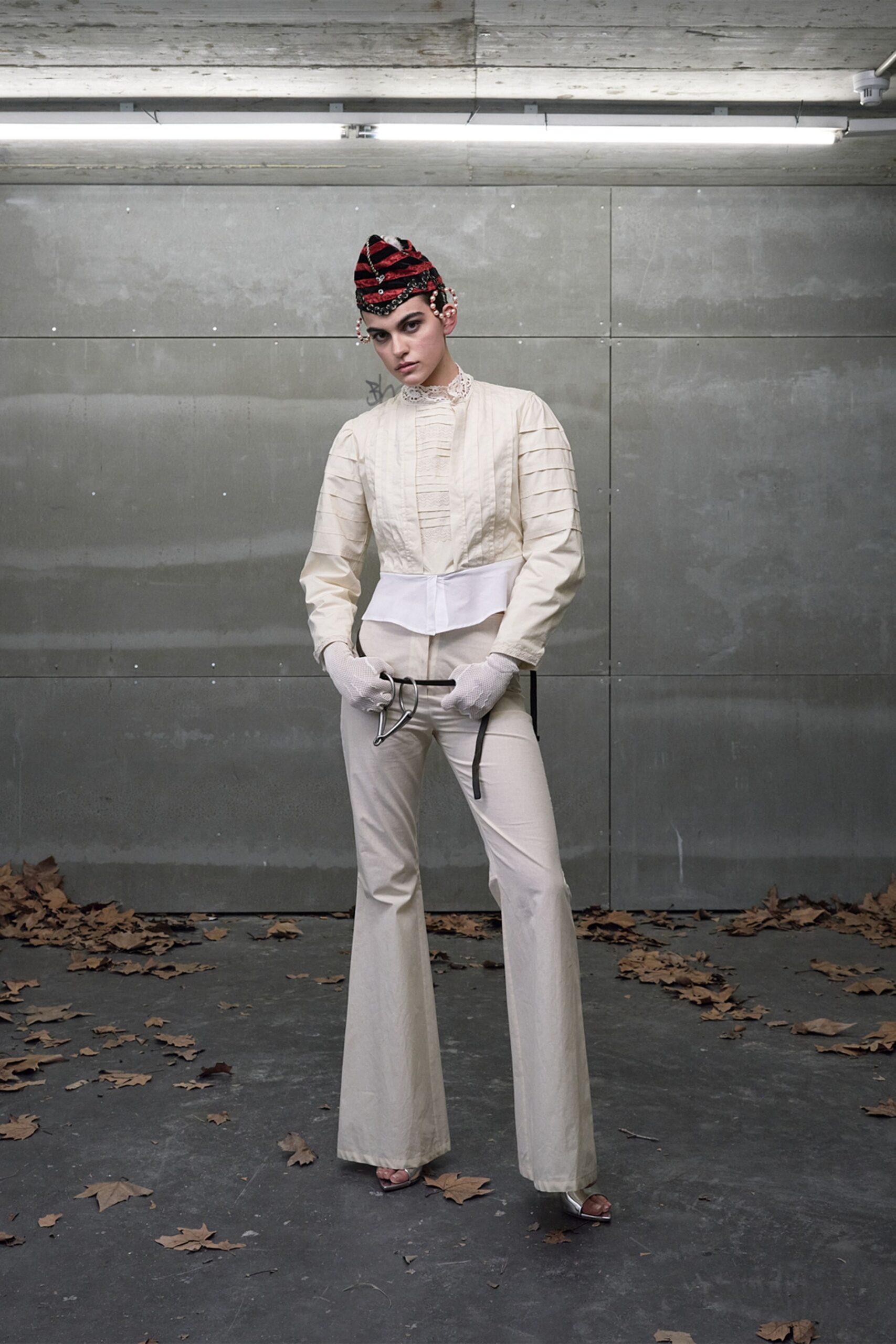 Red and black velvet cap with pearl tassels at the ears. Cream blouse with lace stand collar and pintucked bib. White fishnet gloves. High-waisted cream flares breaking wide over the foot. Silver open-toe heels. Waiting by the freight entrance after a wedding nobody admits attending.