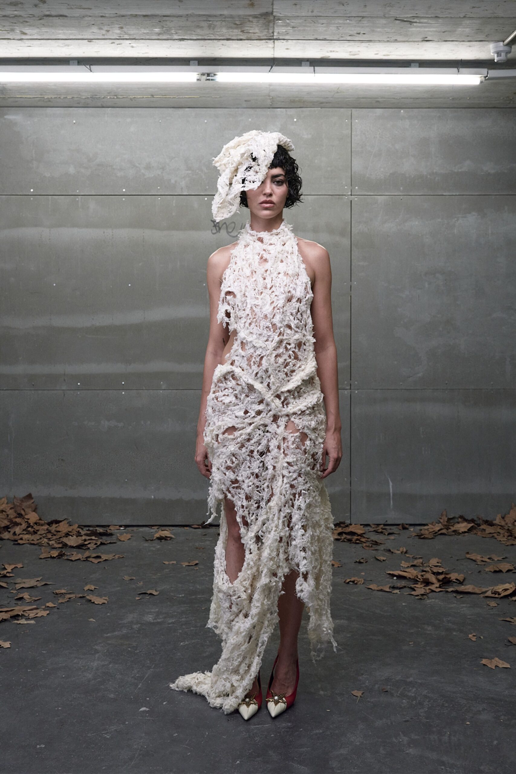 Off-white shredded lace cap slumped over one eye. Openwork ivory dress, matted strands knotted into a trailing uneven hem. Red pumps with ivory pointed caps and jewel clusters. Under garage strip lights, leaves blown in before anyone shut the door.