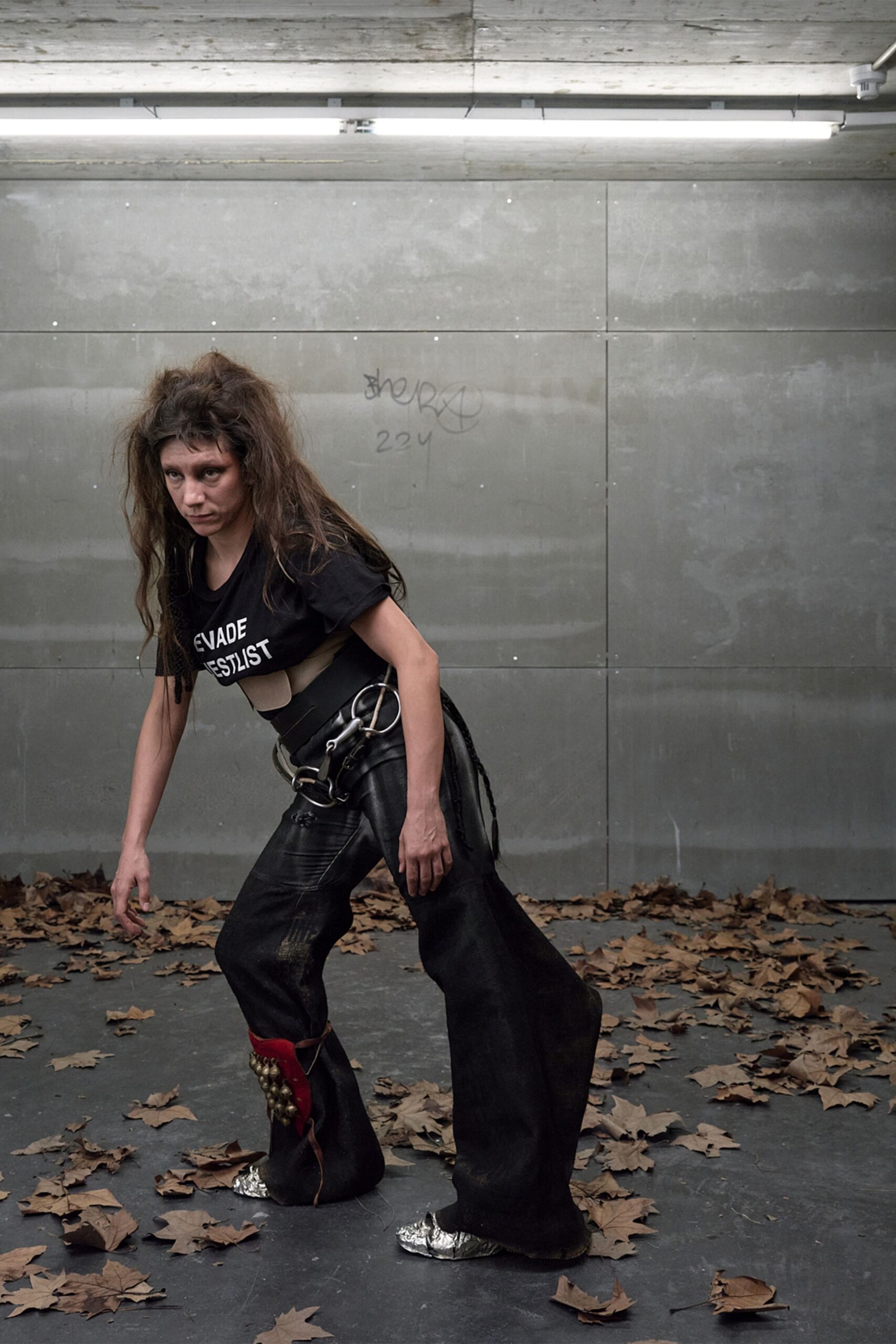 Black slogan tee hitched up to bare one side of the ribcage. Wide black belt harness hung with oversized steel rings and clips. Black coated flares, one leg split by a red inset studded with bells. Crushed silver foil shoes peeking from the pooling hems. Underpass after rain, leaves skittering around the last bus home.