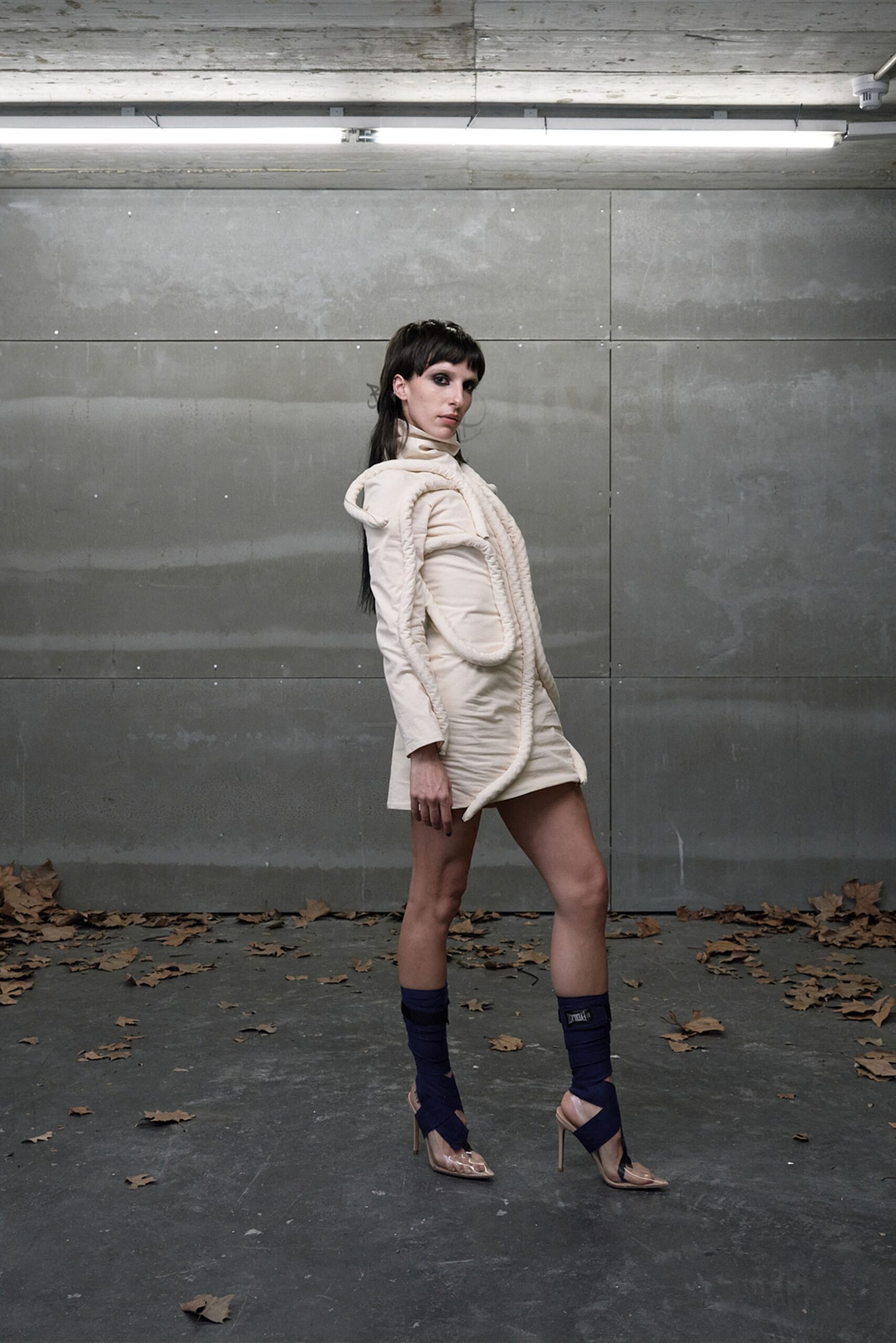 Ivory satin mini dress with a high neck and ruched side seams. Thick tubular trim looping over the shoulders and snaking past the hem. Ink knit leg warmers wrapped tight above the ankles. Clear pointed pumps with nude slingbacks and slim stilettos. Under strip lights after rain, leaves blown into the loading bay.