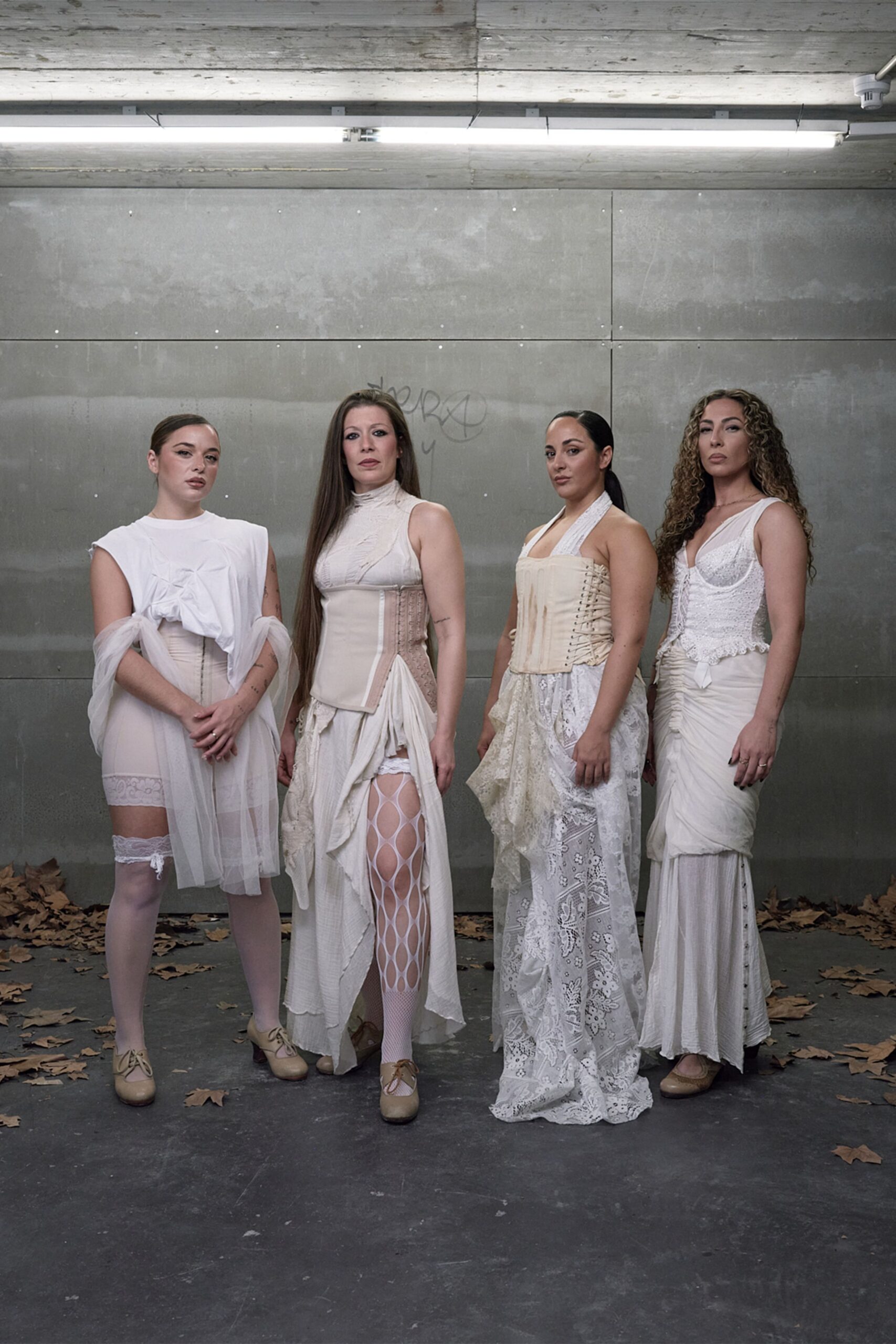 Ivory lace-up corset dress with mixed lace panels and a ruched hip drape. Beige lace-up heels with rounded toes. Under strip lights in a concrete underpass, dry leaves skittering at their feet.