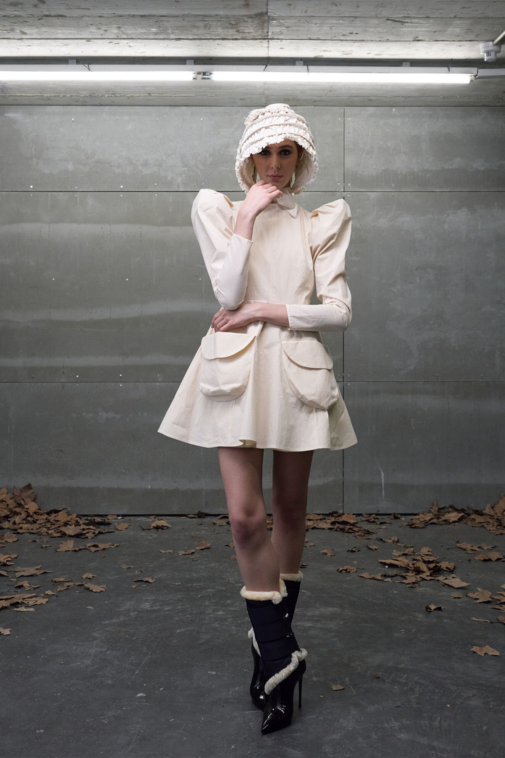 Cream boucle cloche, brim drooping in soft concentric ridges. Ivory mini dress with puffed sleeves and oversized flap pockets at the hip. Black patent stiletto boots wrapped with cream shearling bands. Under strip lights after rain, leaves tracked into the parking garage.