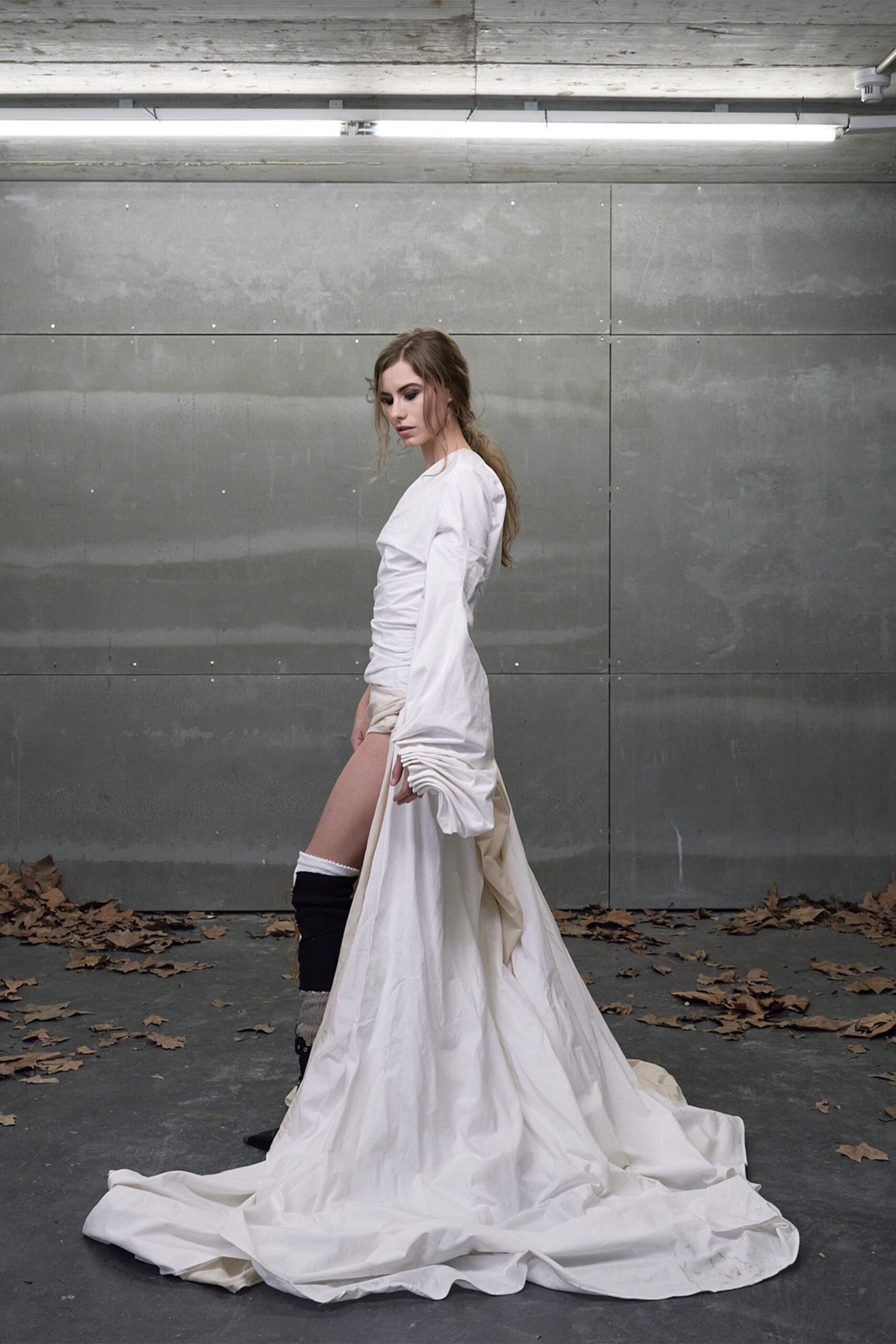 Ivory cotton dress, ruched tight through the torso with a sweeping train. Extra-long sleeves bunched into heavy folds over the hands. Black ribbed leg warmers, white cuff peeking at the knee. Black pointed shoes barely visible under the pooled hem. Under strip lights after rain, leaves blown into the loading bay.