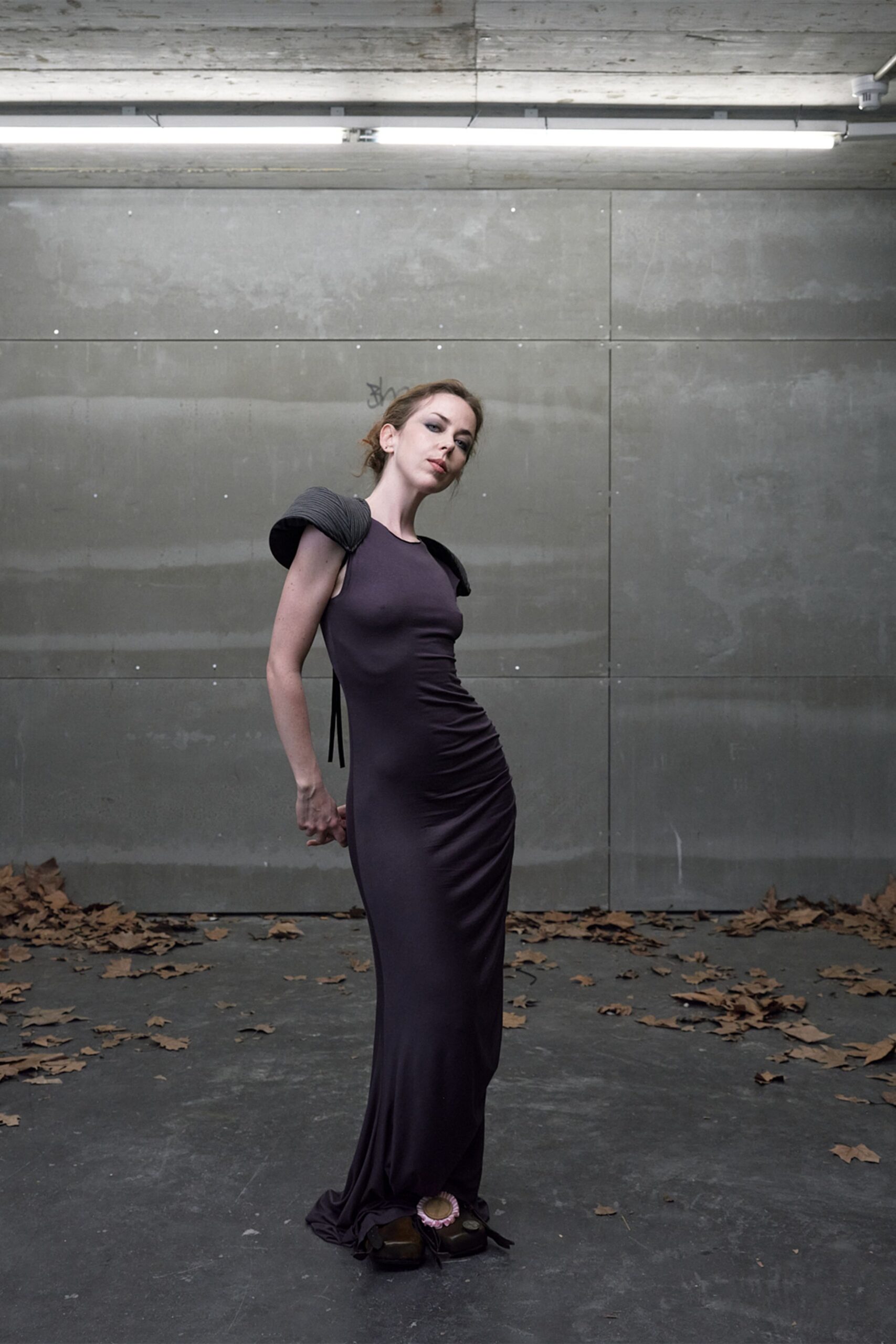Ink-plum jersey column gown, ruched through the hip with a pooling hem. Charcoal ribbed shoulder rolls jutting outward, black ties hanging behind. Square-toe wooden platforms with pink rosette clips. Under strip lights in a car park after the last leaves blew in.