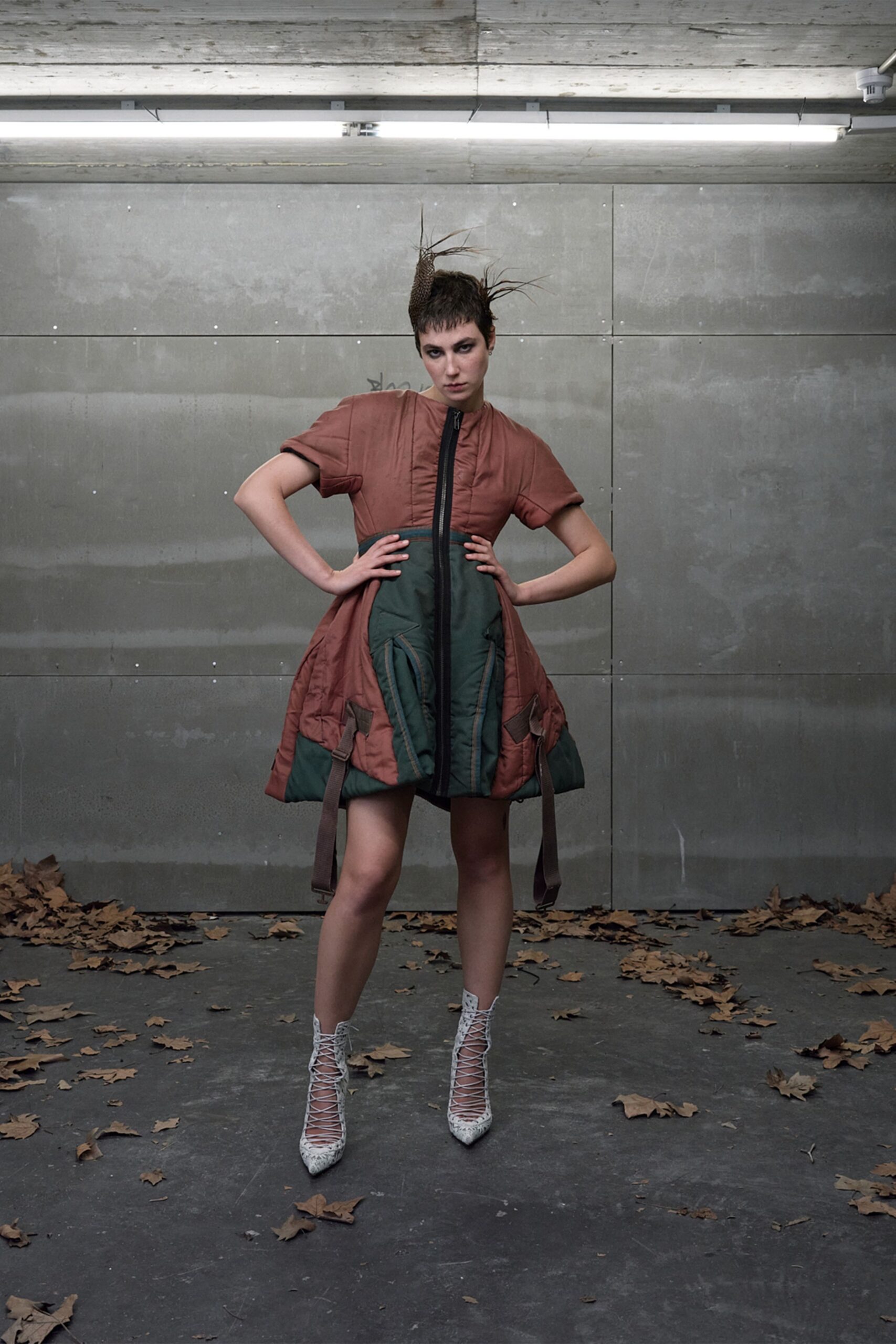 Mesh cone headpiece with wiry quills jutting off the crown. Russet padded dress, black center zip and dark green quilted apron panels. White lace-up ankle boots in a cracked snakeskin finish. Under strip lights in a parking garage, leaves blown against the wall.