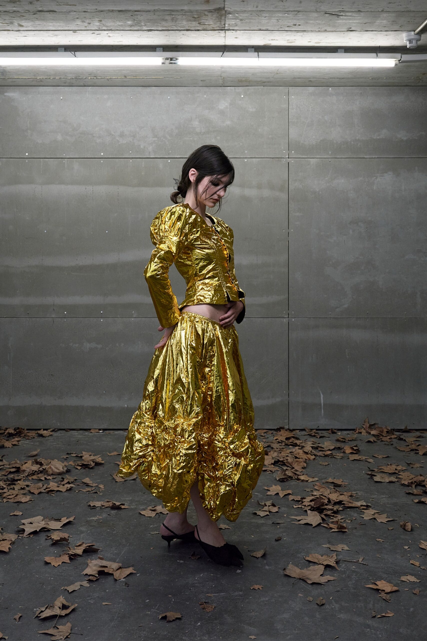 Crinkled gold lamé jacket, puff shoulders and tiny hook closures. Matching drop-waist skirt, gathered into a swollen bubble hem. Black pointed mules with a low curved heel. Under strip lights in a concrete garage, leaves skidding in at the door.