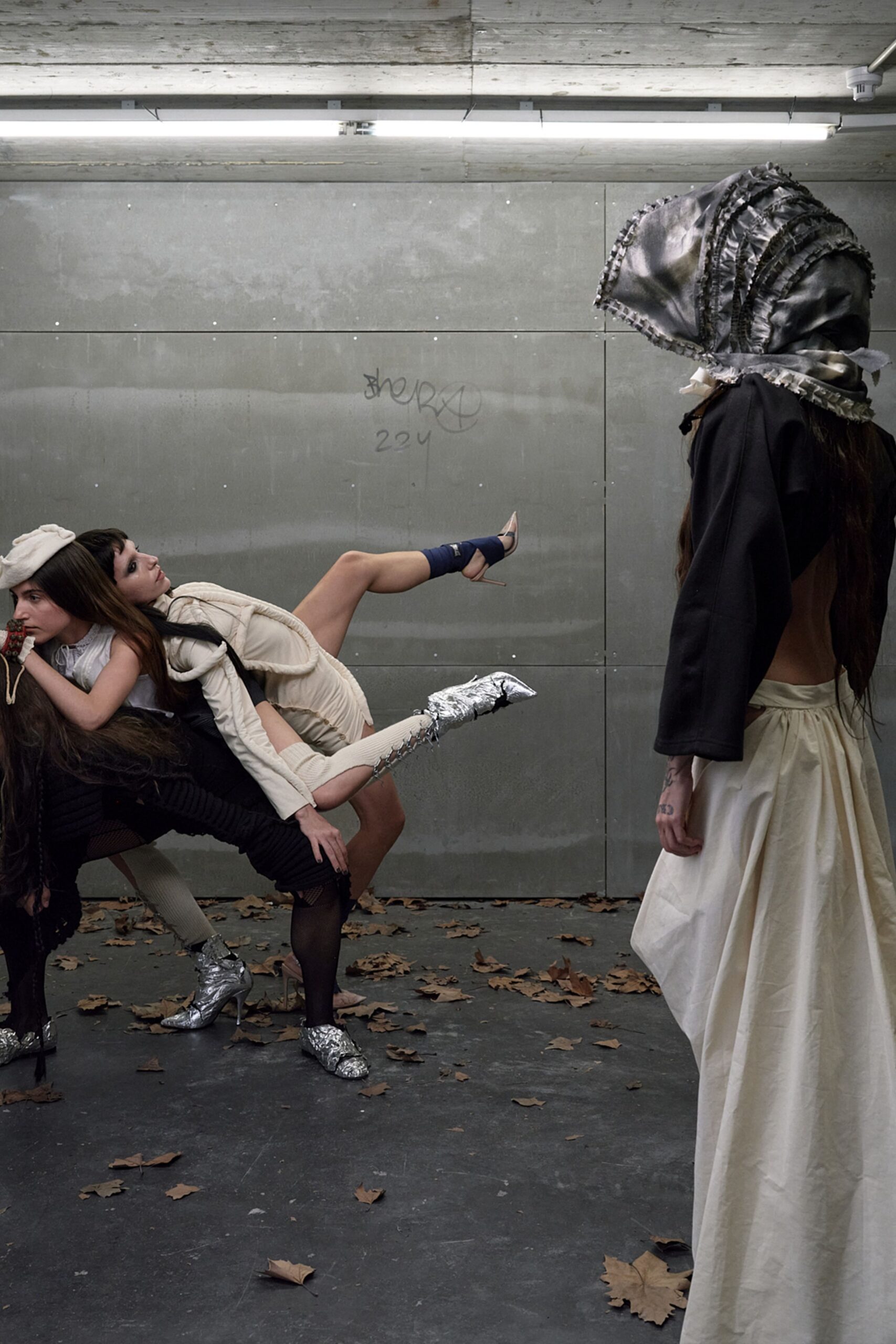 Oversized black satin bonnet ringed with frilled bands and tied under the chin. Black capelet cut broad at the shoulder, hanging stiff over bare back. Full ivory cotton skirt, deeply pleated and swelling from a low tie waist. Under strip lights in a concrete underpass, leaves dragged in from the street.