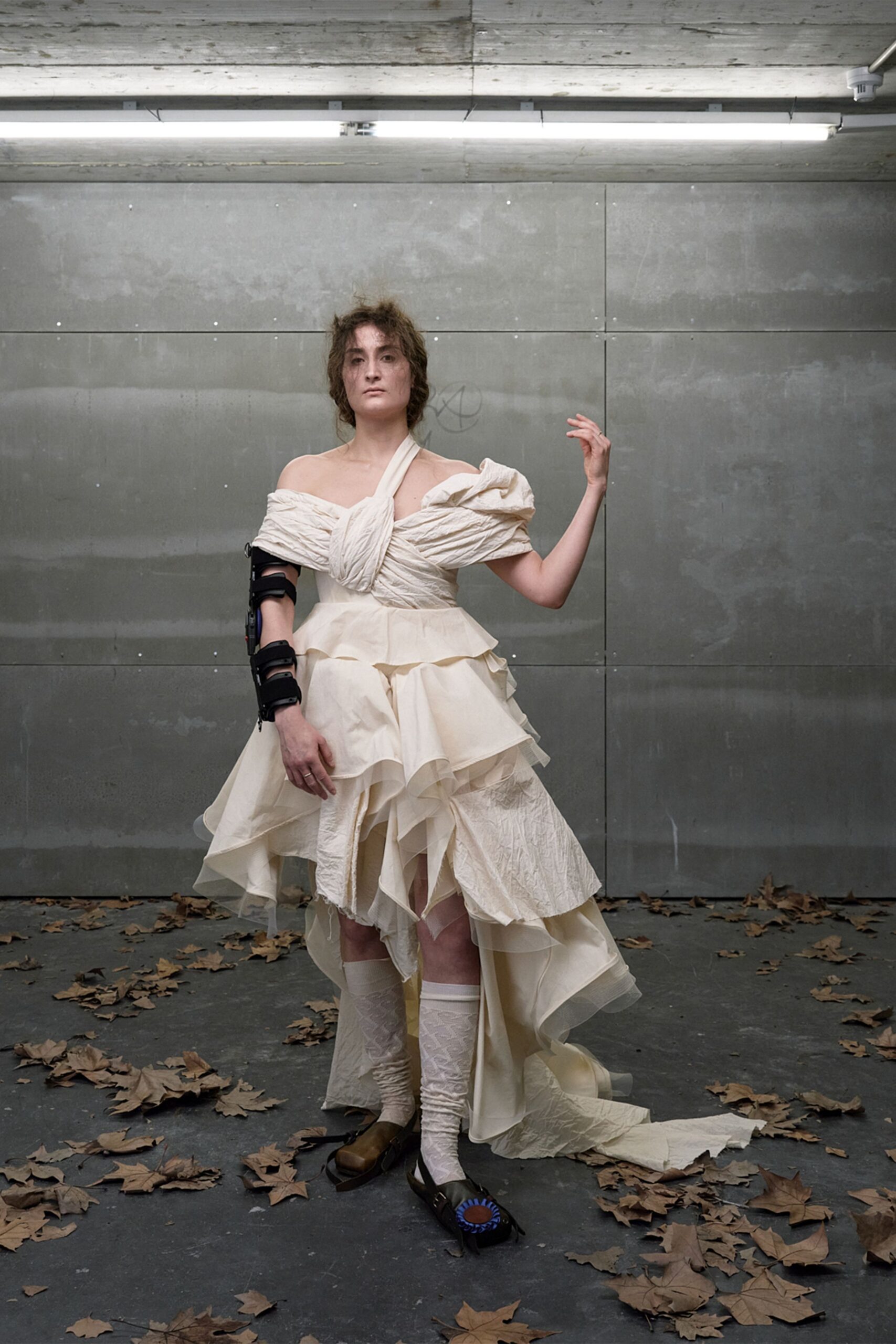 Ivory dress with ruched off-shoulder bodice and cascading raw-edged organza tiers. Black orthopedic brace strapped from upper arm to wrist. Cream knit leg warmers, softly slouched and semi-sheer. Square-toe black flats, one topped with a blue rosette. Leaves blown into an underpass after the courthouse wedding.