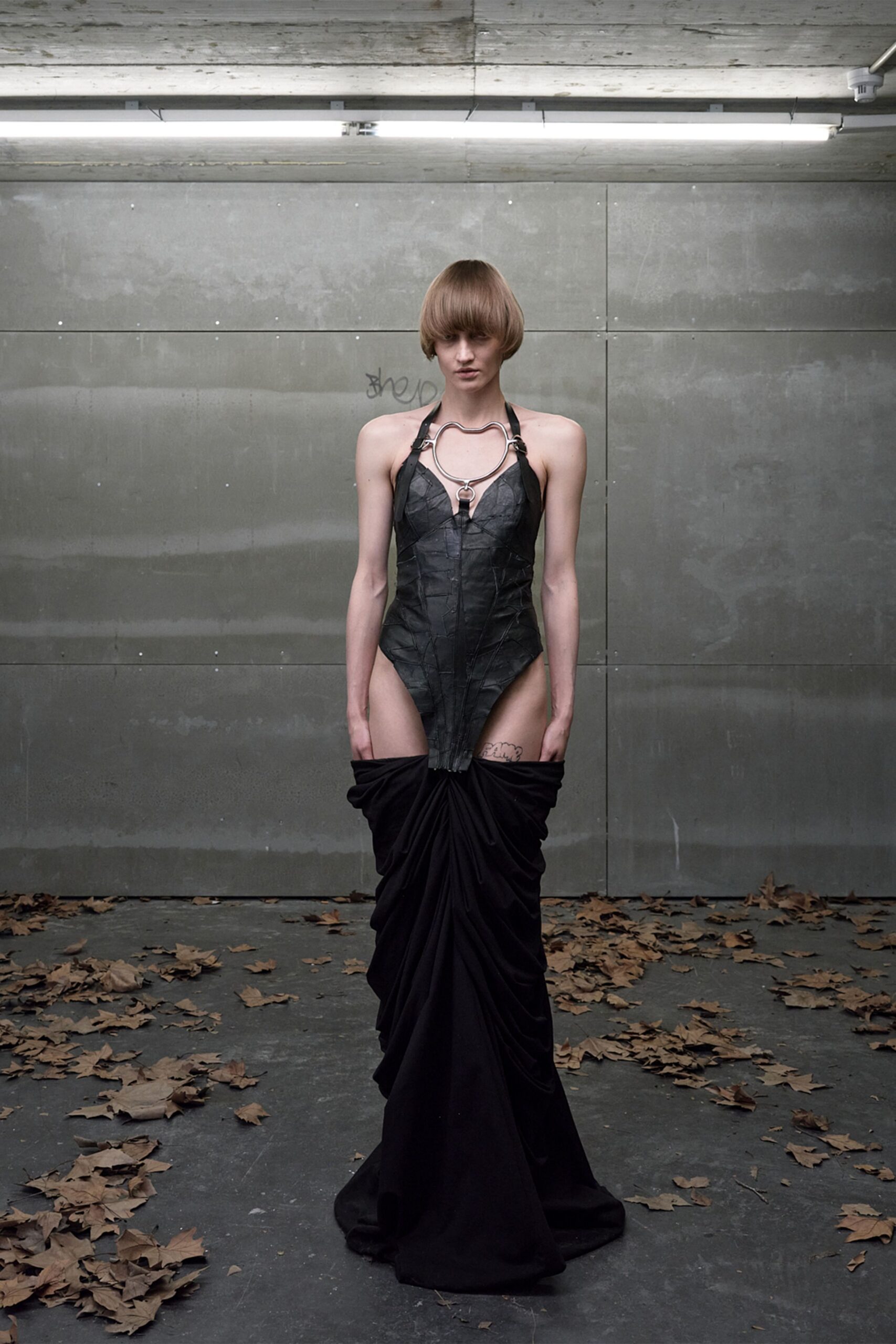 Black pieced leather halter bodice, wrinkled like pressed leaves. Large silver collar frame with an O-ring dropping at the sternum. Black jersey skirt ruched hard at the hips, pooling into a train. Under strip lights in a courtyard gone cold, leaves skittering at your feet.