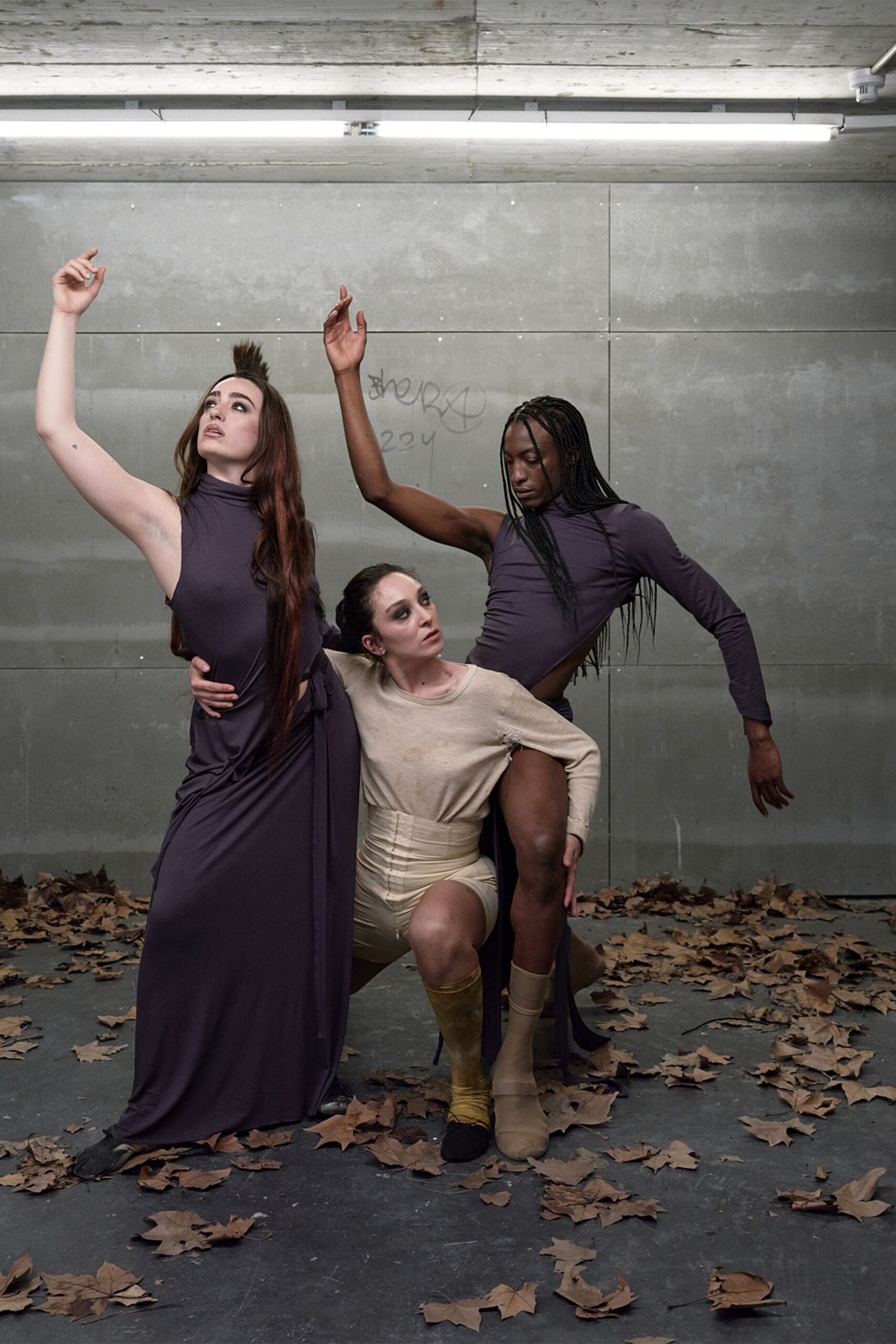 Plum jersey dress with a high neck and one bare arm. Self-tie sash knotted low, one long end falling to the floor. Black split-toe flats peeking from the pooled hem. Dry leaves blown into an underpass before the afternoon dance call.