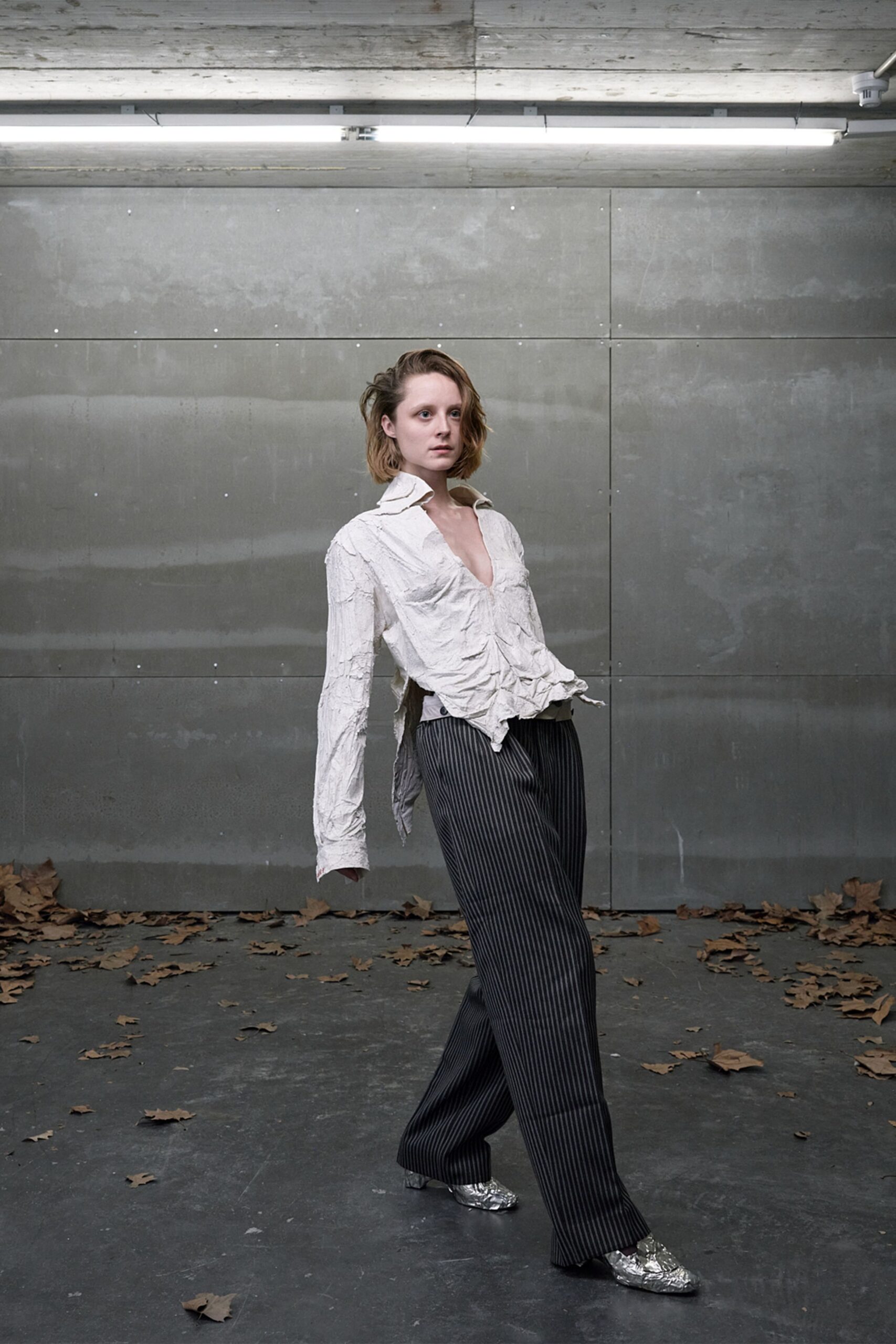 Crushed ivory shirt, collar kicked up and hem left in ragged points. Charcoal pinstripe wide-legs with a pale waistband peeking above. Silver crinkled loafers catching the striplight. Walking through an empty loading bay with leaves skittering indoors.