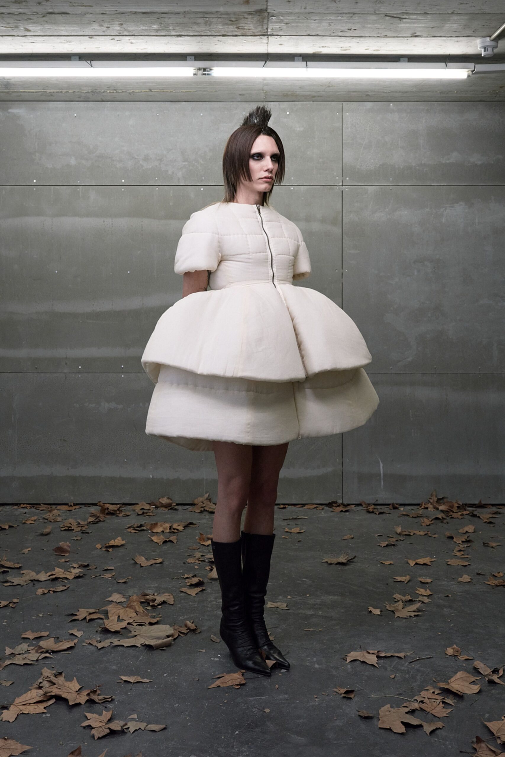 Ivory quilted dress with puff sleeves and two stuffed hoop tiers. Black leather boots, pointed toes peeking beneath the padded hem. Windblown leaves skittering through a concrete underpass at dusk.
