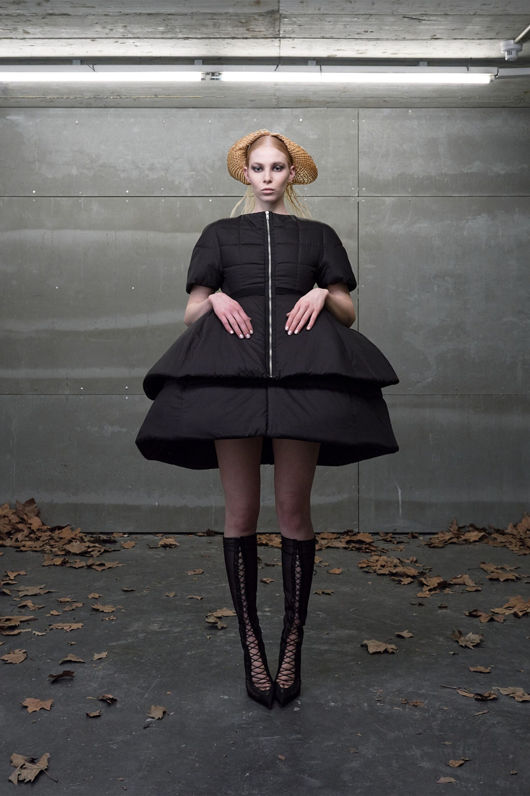 Open-weave straw hat splayed into two side horns. Black padded dress, center zip and tiered pannier-like bubble skirt. Black lace-up mesh boots with razor-point toes. Under strip lights after rain, leaves blown into the loading bay.