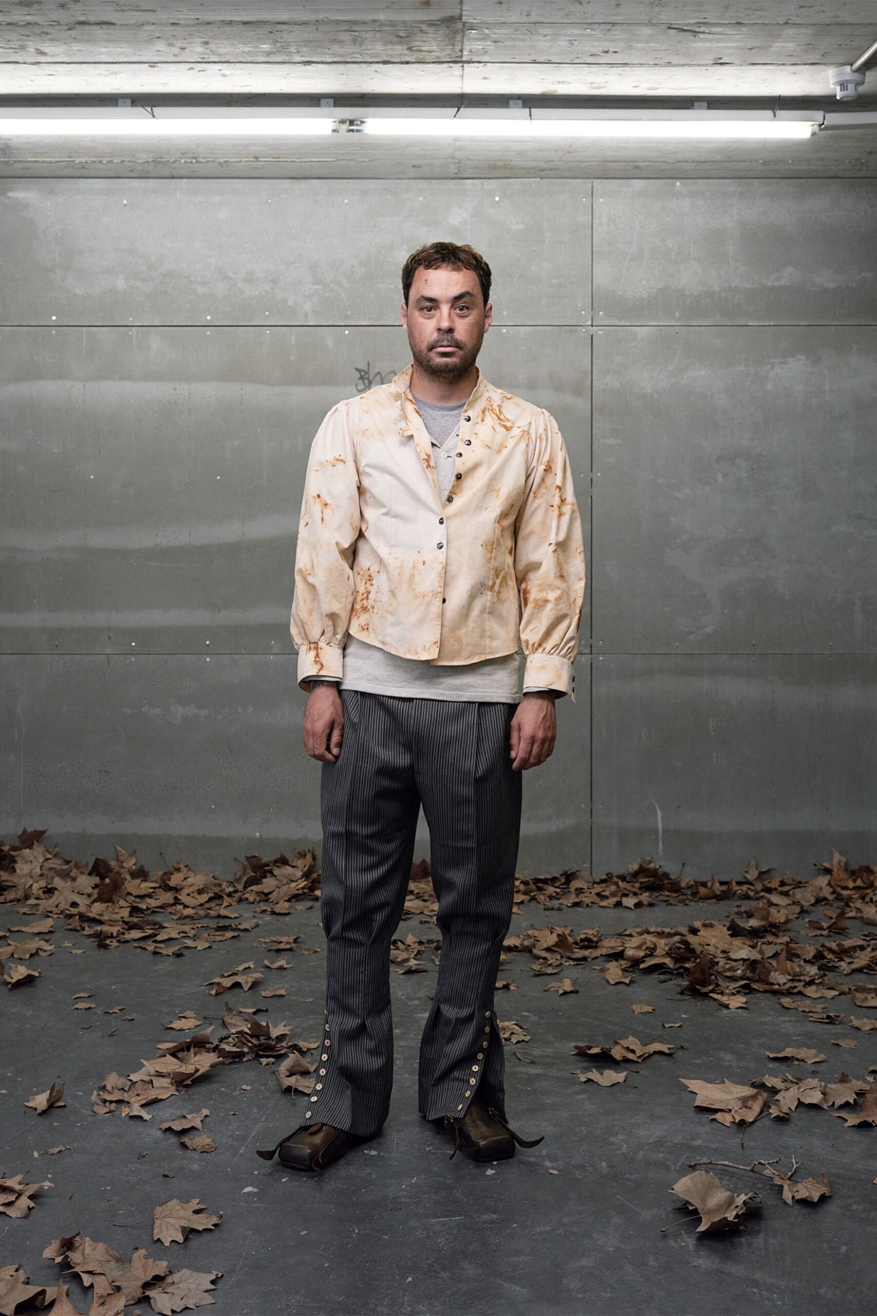 Cream poplin shirt washed with rust-like brown stains, black snaps askew. Grey jersey tee showing at the deep open placket. Charcoal pinstriped trousers split at the hem with pearl buttons. Square-toe leather shoes with long curled tongues. Under strip lights after sweeping leaves in from the loading bay.