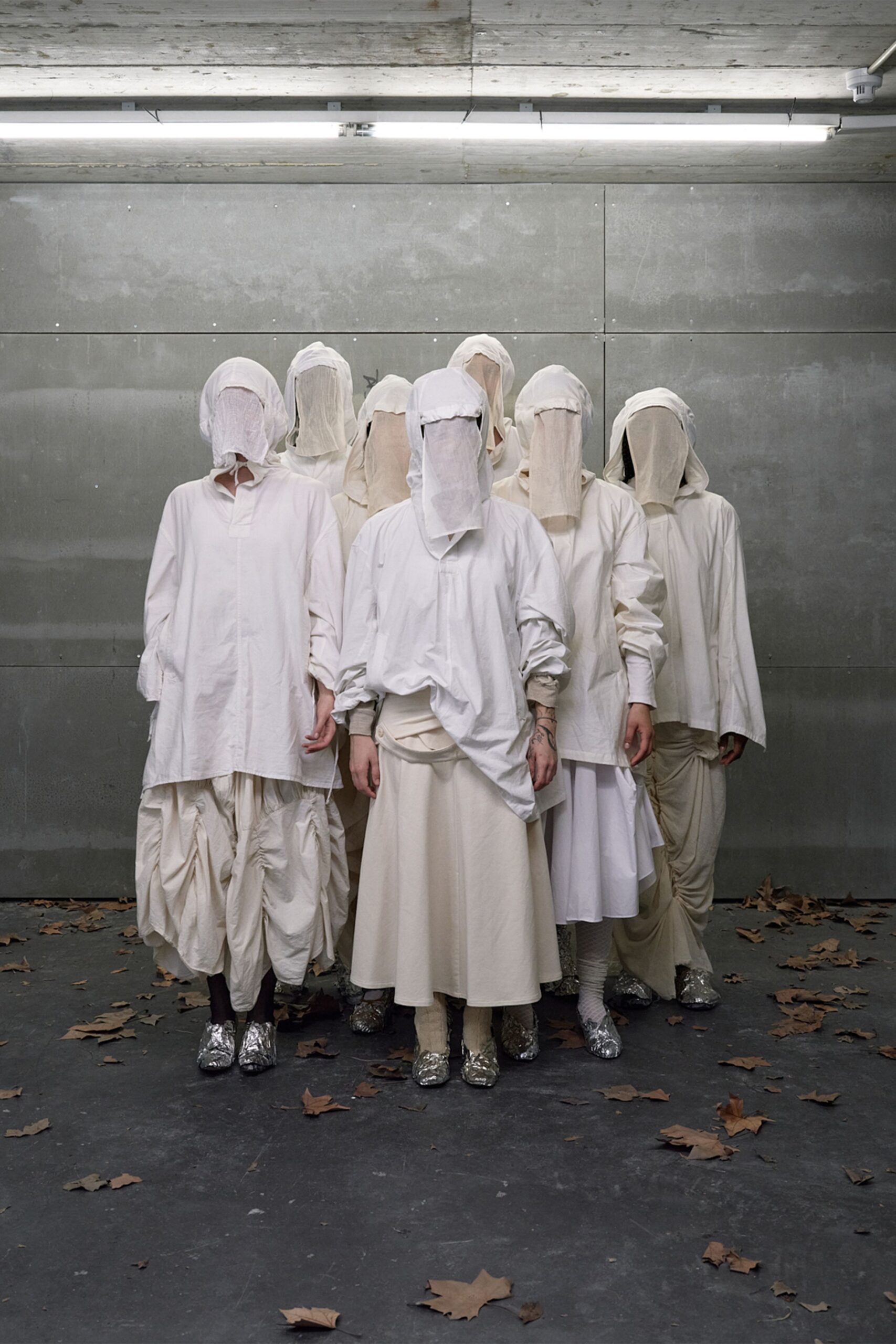 Gauzy white hood dropping into a full face veil. Oversized white cotton tunic, placket half hidden by the drape. Milk-white skirt falling in a plain heavy column. Silver foil boots, creased hard across the vamp. Under strip lights in a concrete loading bay, leaves blown in after dusk.