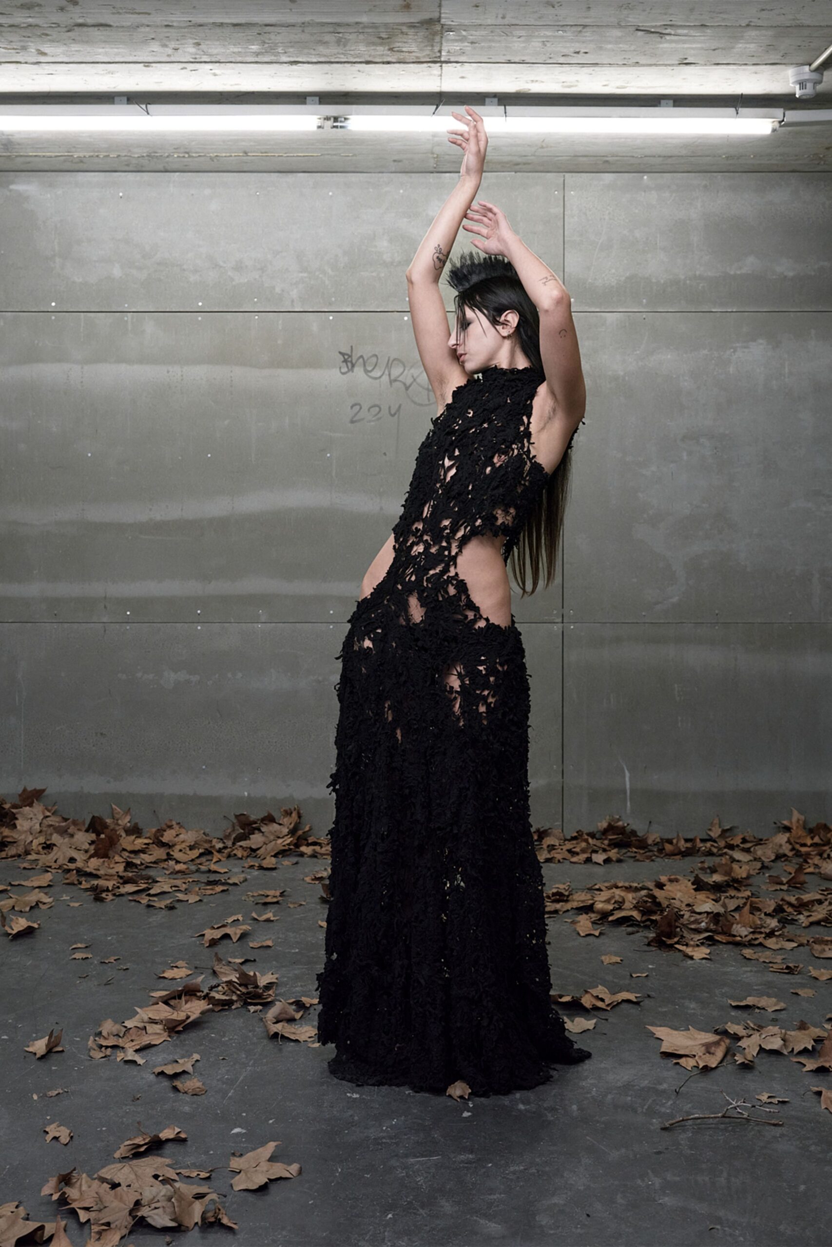 Black shredded openwork gown with a high neck and bare side cutouts. Underpass concrete after rain, leaves skittering under the strip lights.