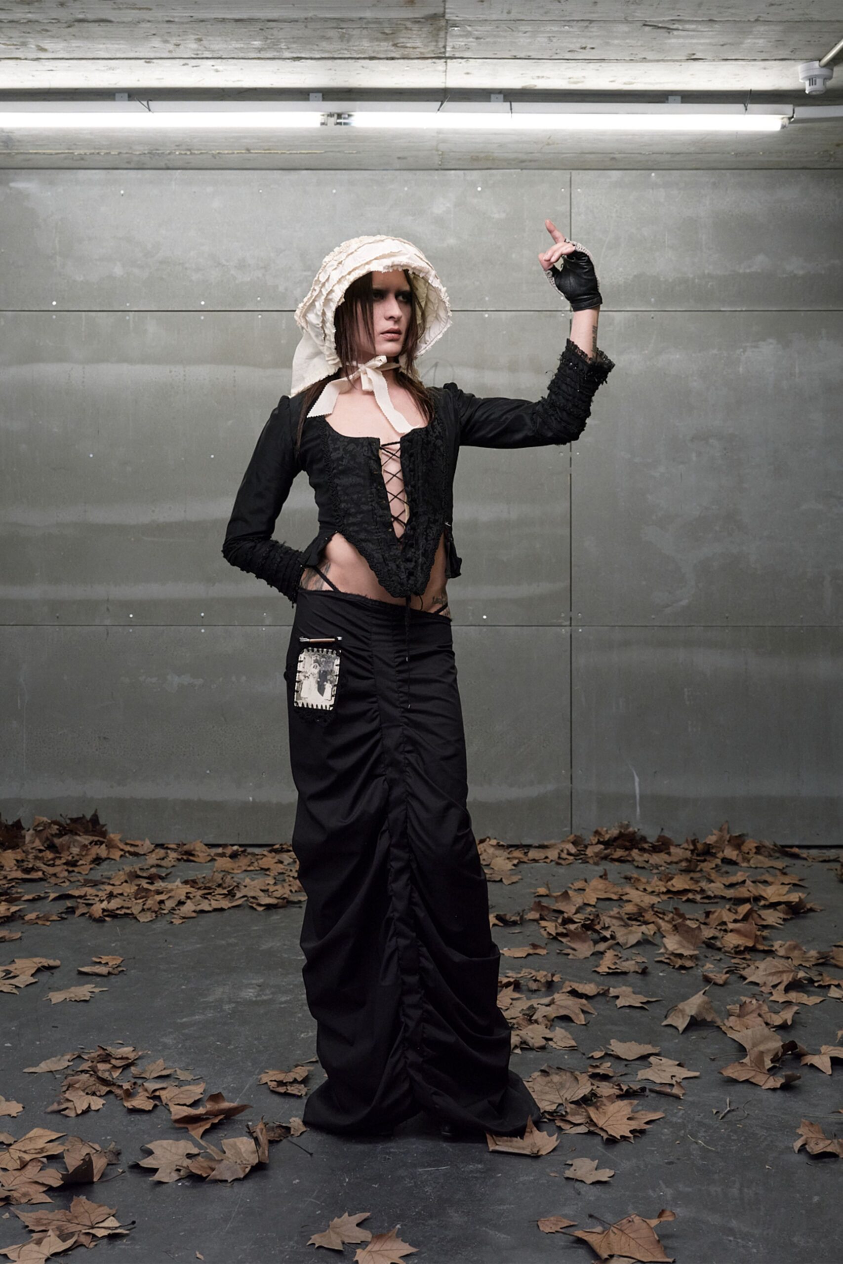 Ivory bonnet stacked with raw ruffles and tied under the chin. Black lace-up top with a plunging split front and ruched sleeves. Black fingerless leather glove. Floor-length black skirt, center seam tightly ruched, patch pocket hanging low. Walking through a concrete underpass after the trees have dropped everything.