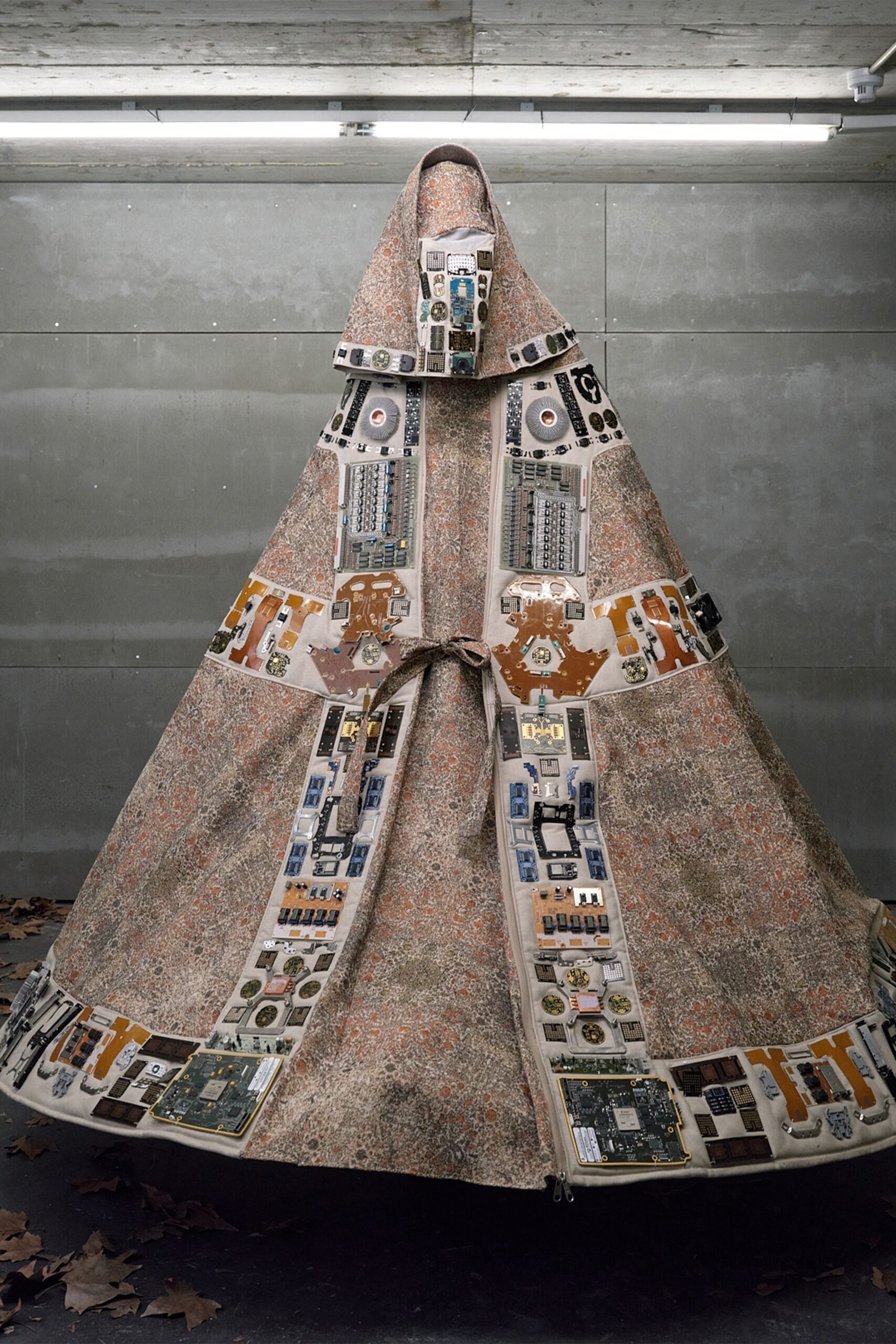 Floor-length mottled cloak appliqued with salvaged circuit boards and disk drives. Matching speckled textile pulled smooth across the whole face. Narrow self-fabric cord knotted loosely at the center opening. Under strip lights in a concrete garage, dressed for the server room's funeral.