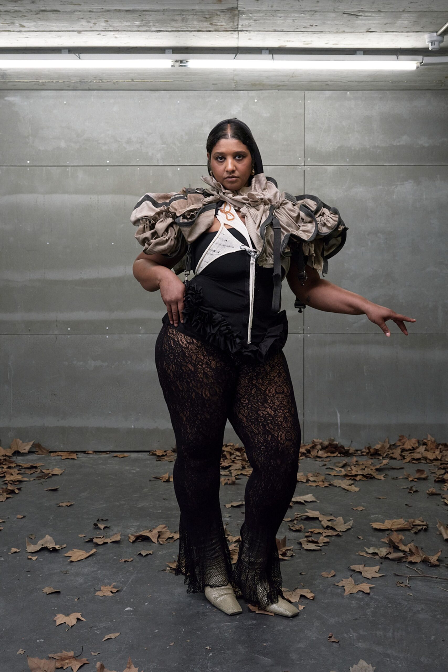 Taupe ribbed hooded shrug, gathered into bulbous ruffles with black binding. Black bodysuit with a white curved zip panel and ruffled peplum. Black floral lace stockings, hems loosening into a fishnet flare. Pale pointed flats in cracked metallic leather. Under strip lights while dry leaves blow through the loading bay.