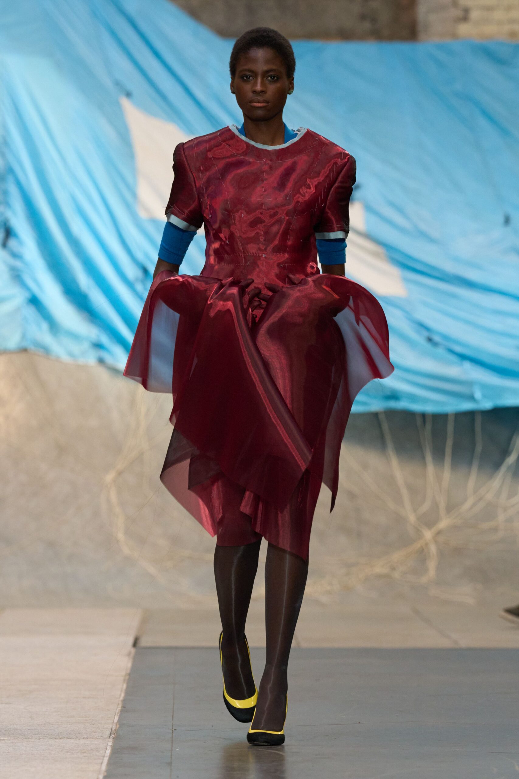Burgundy translucent organza dress with padded shoulders and a ballooned overskirt. Electric-blue jersey undersleeves, cut off bluntly below the short sleeves. Glossy black tights. Black pumps edged in acid-yellow at the vamp. Crossing a windblown fairground after the plastic sheeting came loose.