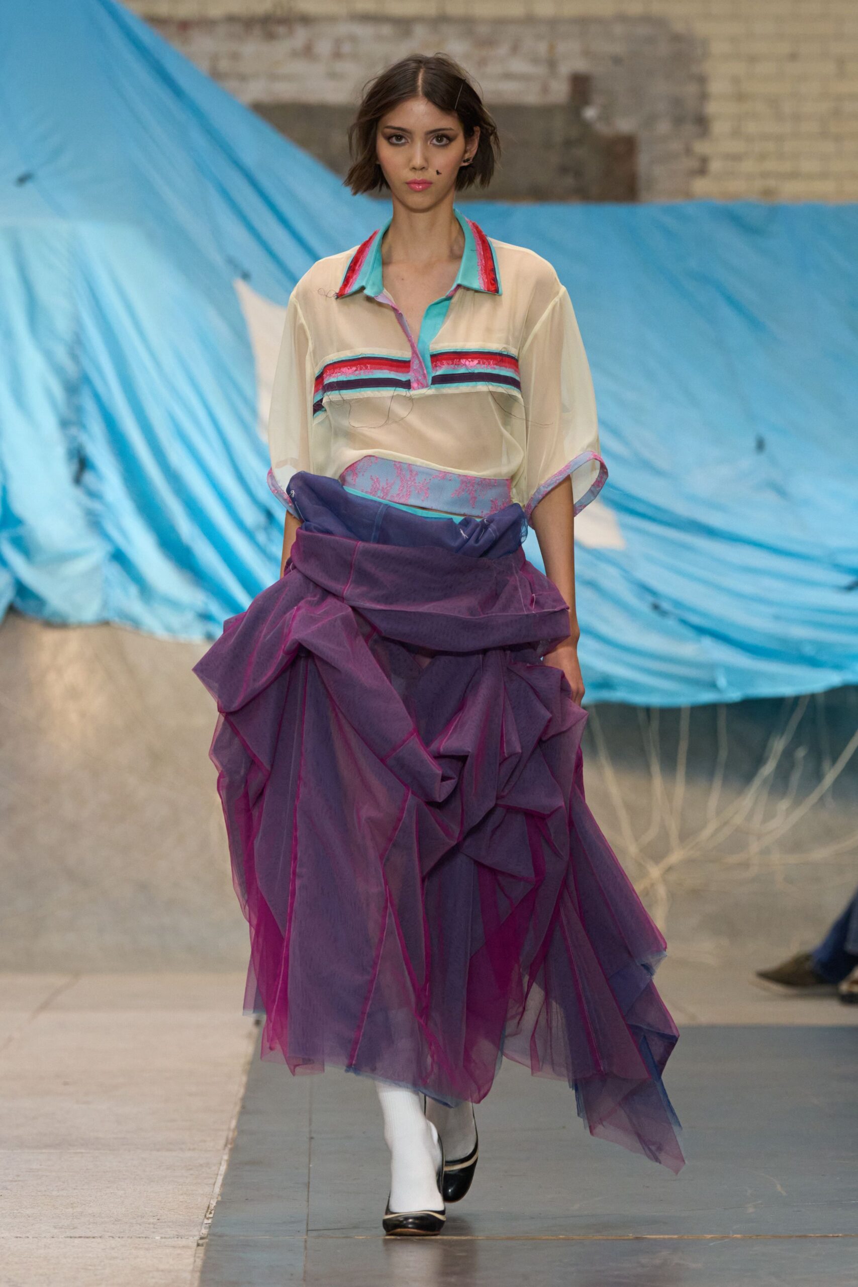 Sheer cream blouse with turquoise placket and striped bands across the chest. Voluminous plum organza skirt, wrapped high and collapsing into angular ruffles. Opaque white tights. Black cap-toe pumps with a narrow white rim. Wind off the fairground, tarpaulins snapping over half-packed stalls.