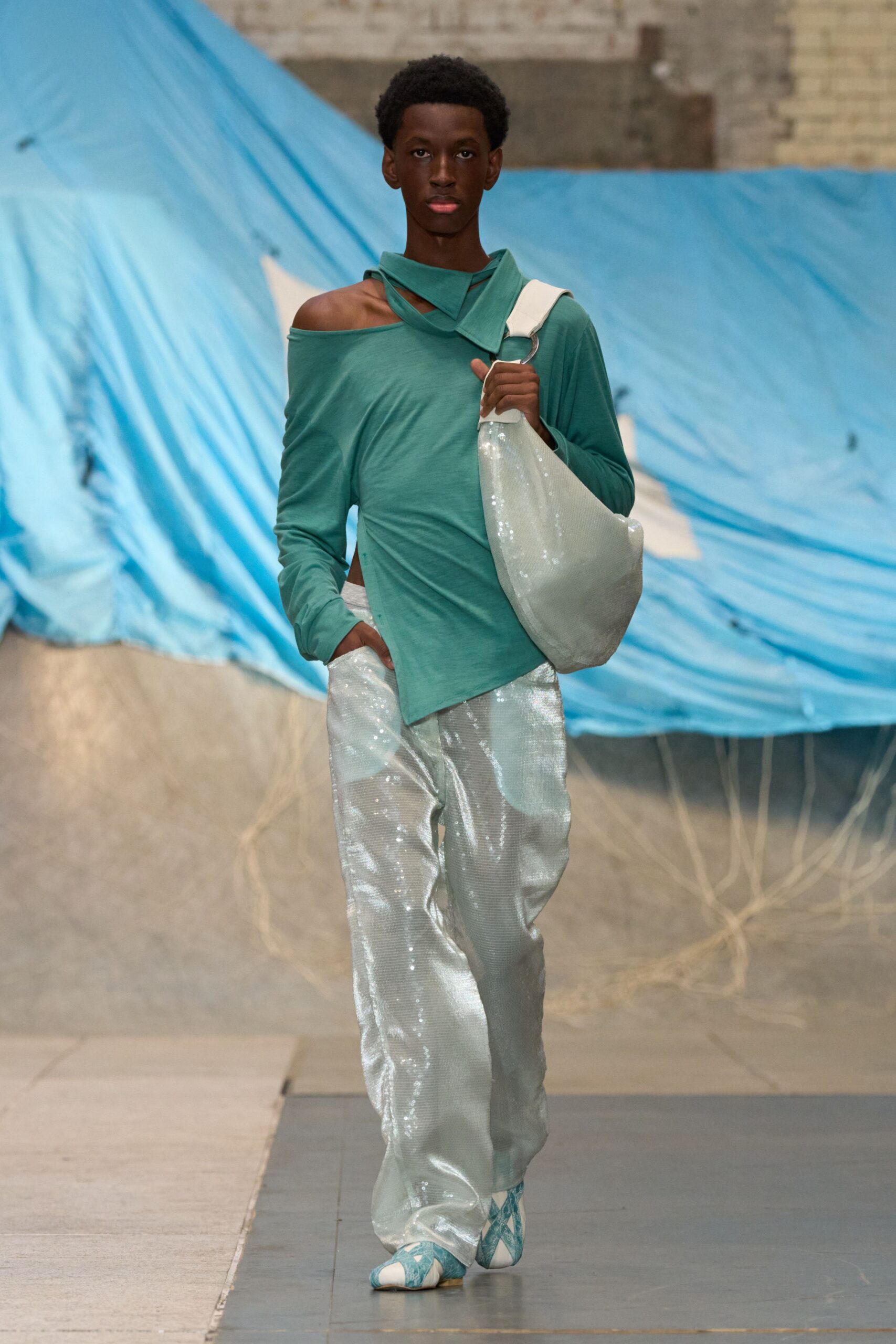 Seafoam jersey top draped off one shoulder with a crossed funnel neck. Slouchy white sequined shoulder bag on a thick strap and metal ring. Loose silver sequined trousers pooling softly over the instep. Pale blue argyle flats. Walking across a fairground after rain, tarp still snapping in the wind.