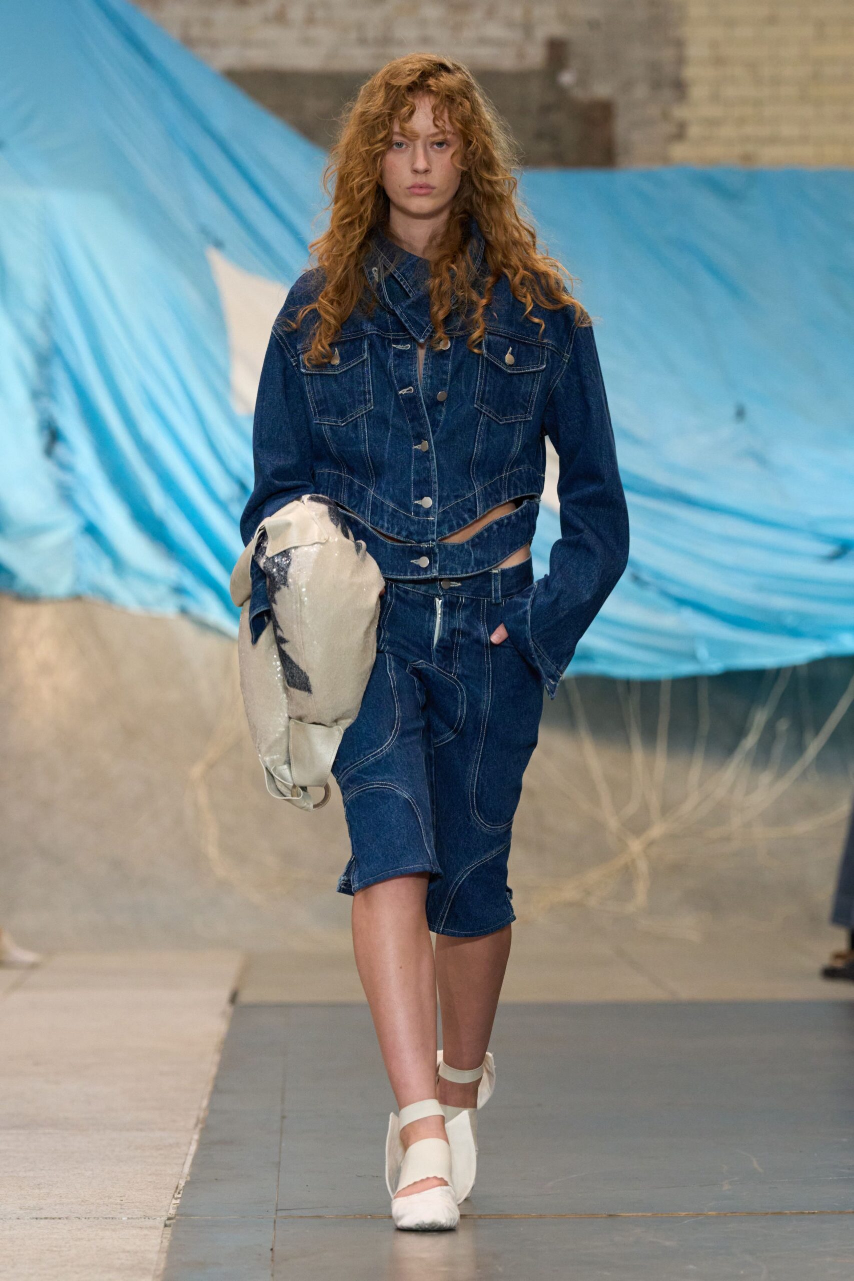 Dark indigo denim jacket, pointed yoke and side cutouts under the hem. Matching denim pencil skirt with looping contrast topstitching and a front zip. Cream sequined slouch bag, black motif half-hidden in the fold. White split-toe flats with broad elastic straps. Crossing a drafty warehouse with salt grass caught at the doorway.