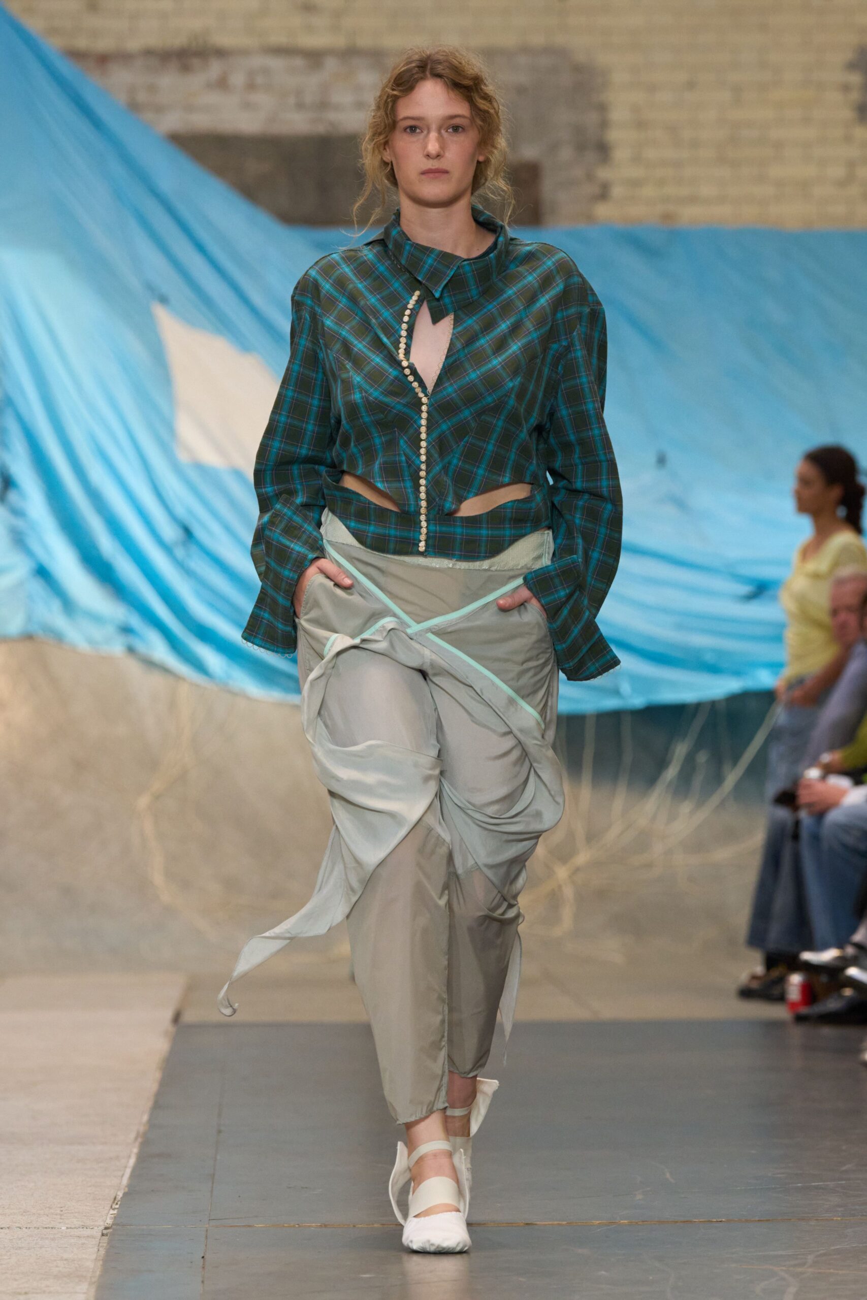 Blue-green tartan blouse cut open at the waist, pearl-buttoned on the slant. Pale grey parachute trousers with a tied overskirt panel and mint binding. White wrap flats, toes softly puckered. Walking across a sports hall after the emergency cots were folded away.