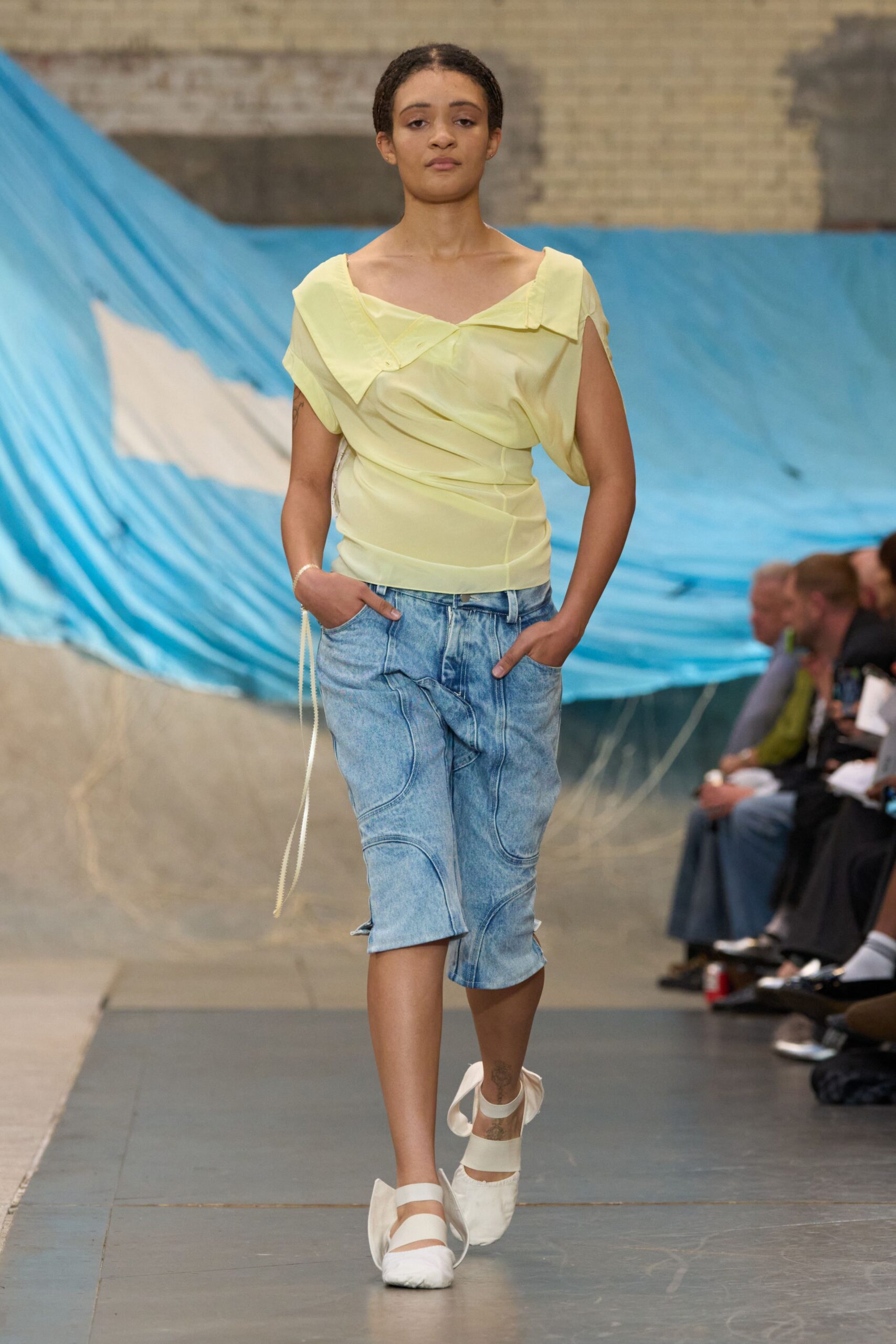 Butter-yellow satin top with a wide folded neckline slipping off one shoulder. Stonewashed denim pencil skirt paneled in looping seams, one flap jutting at the hip. White flats strapped with grosgrain bands and soft bow-like fins. Walking past tarps snapping in the wind at an empty lido.