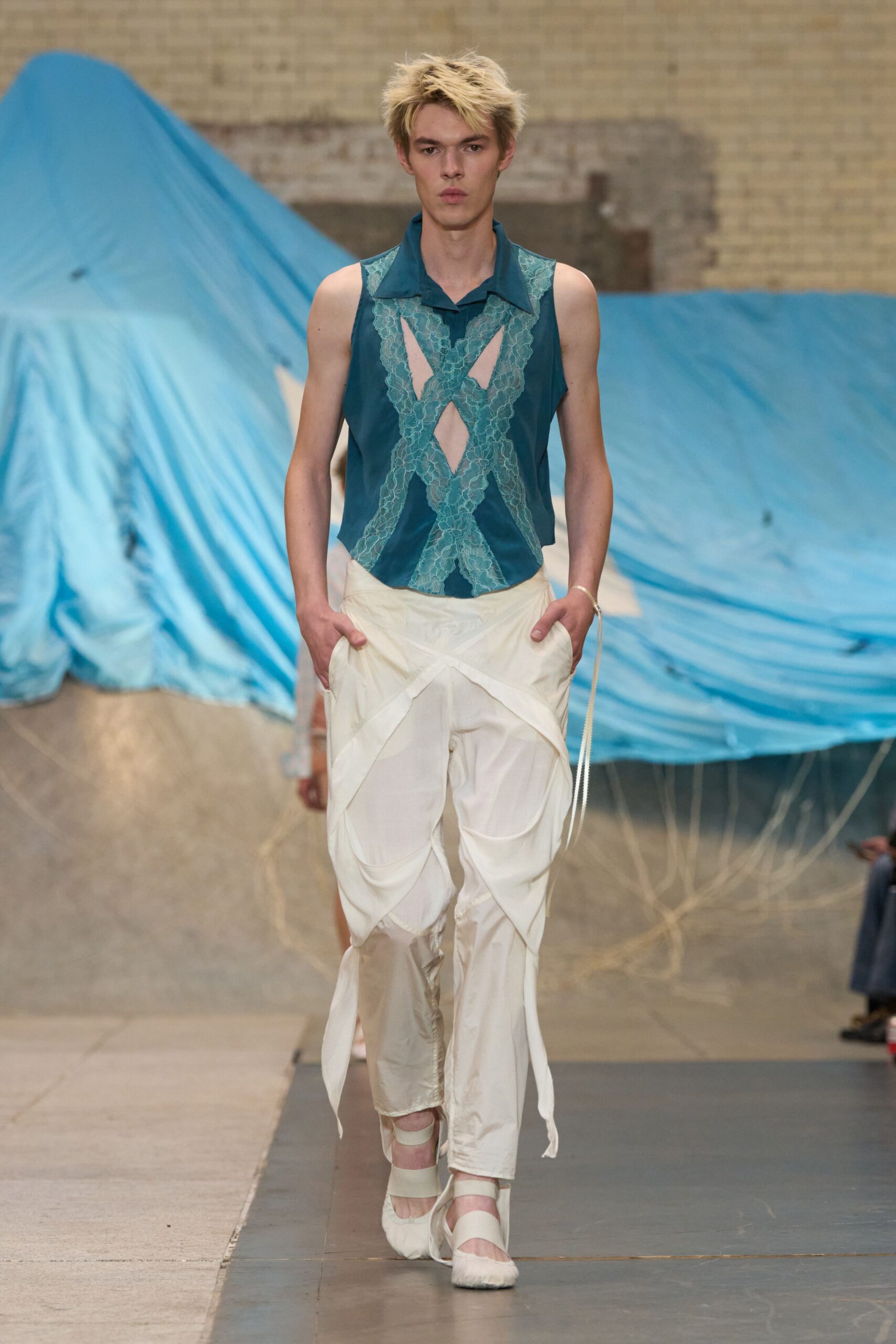 Teal collared blouse with pale lace crossing into raw-edged cutouts. Ivory satin trousers draped in looping panels, one cord end trailing. Cream ballet heels with wide elastic bands over the instep. Walking out of a beach wedding after the champagne went warm.