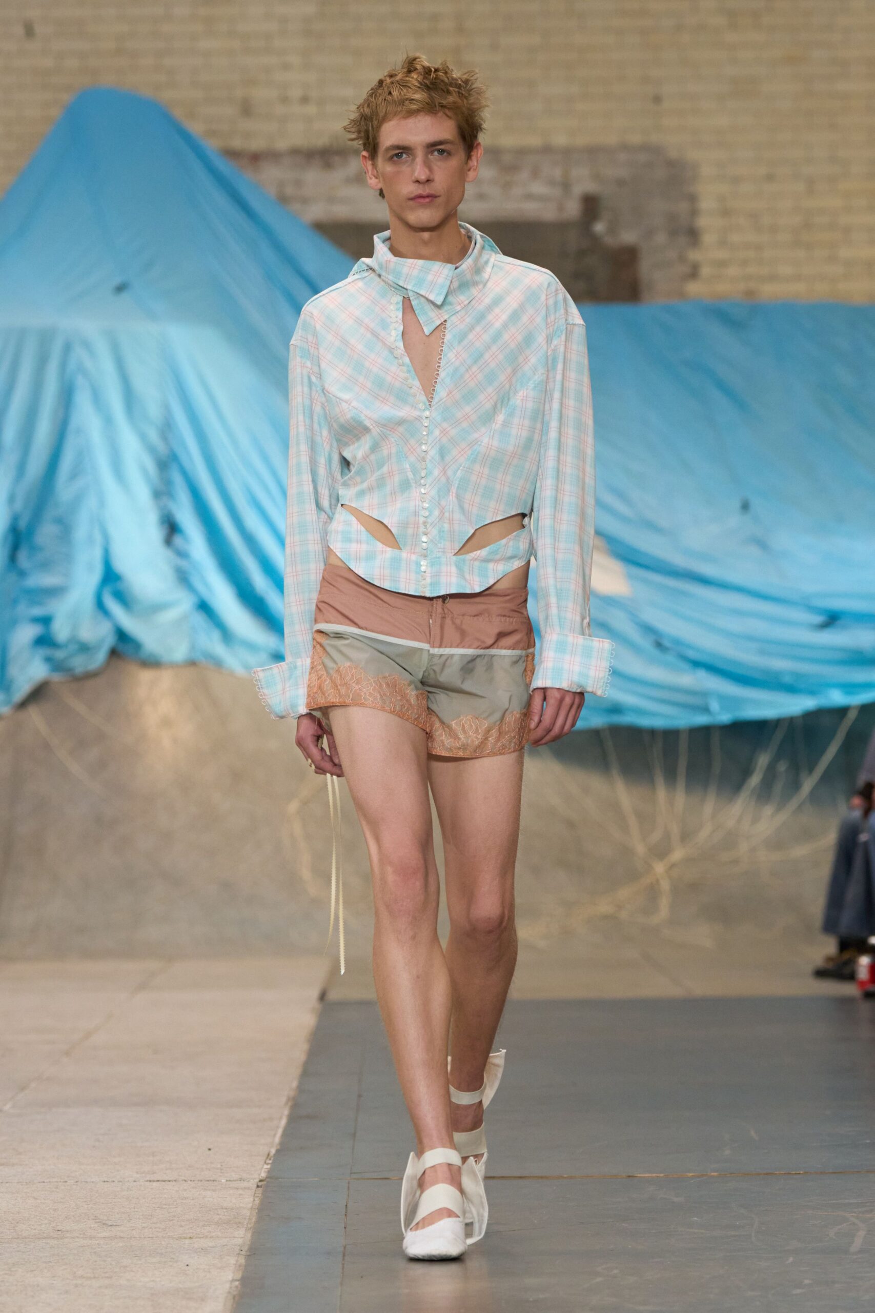 Pastel plaid shirt with side cutouts, pearl-loop placket and one long cord end. Dusty nylon shorts paneled in taupe and sage, peach lace skimming the hem. White slingback flats with broad elastic bands across the instep. Walking across a campsite at dawn before anyone else unzips the tent.