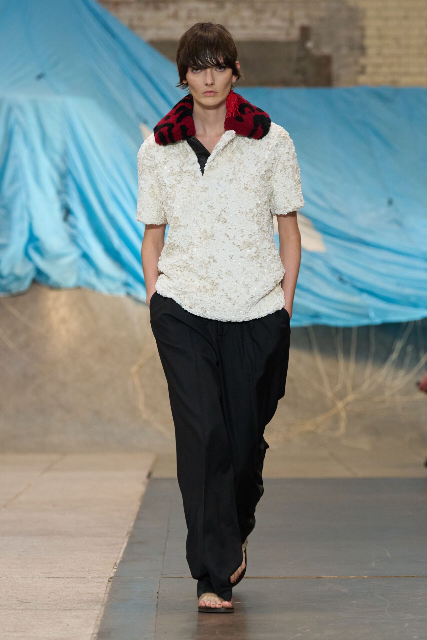 Cream paillette polo, shaggy with a black inset at the split neck. Oversized red-and-black animal-pattern collar in plush pile. Black fluid trousers falling long over the toes. Flat black thong sandals. Walking across a half-finished loft while the painters' tarps are still up.