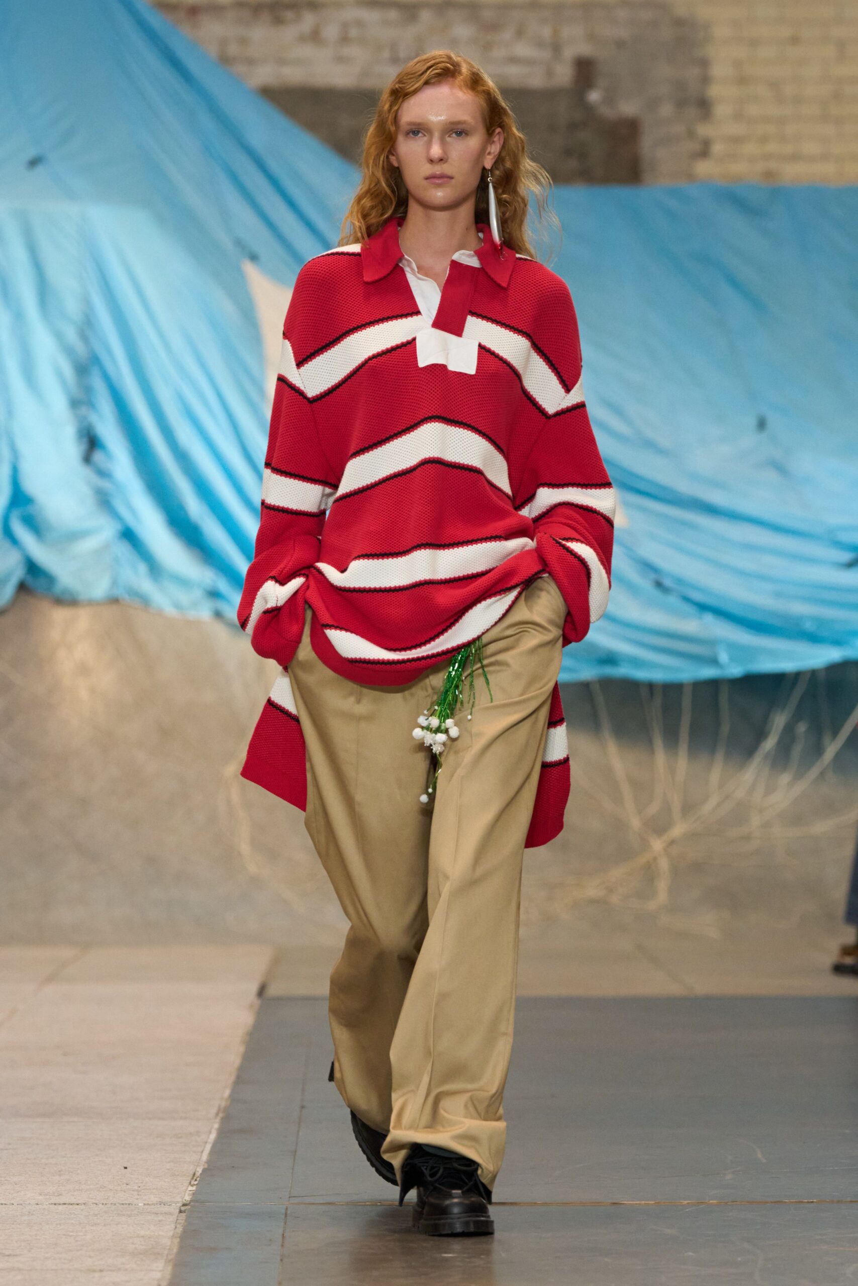 Oversized scarlet waffle-knit rugby with cream chevrons edged in black. Khaki twill trousers cut loose, hem breaking over the shoe. Single long silver drop earring. Black lug-sole boots. Walking across a fairground lot after the tents have come down.