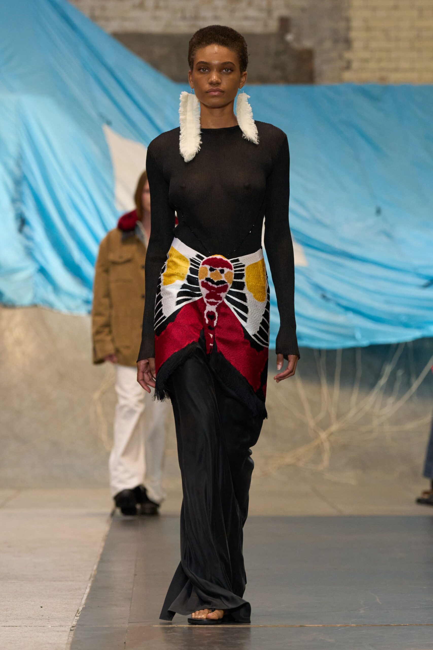 Long white feather earrings brushing the shoulders. Black sheer ribbed knit top, seams curving inward at the waist. Tapestry wrap skirt with a sunburst figure and fringed point front. Black satin trousers pooling over the sandal. Barely there black sandals. Walking past a painted beach tarp after the fair has packed up.