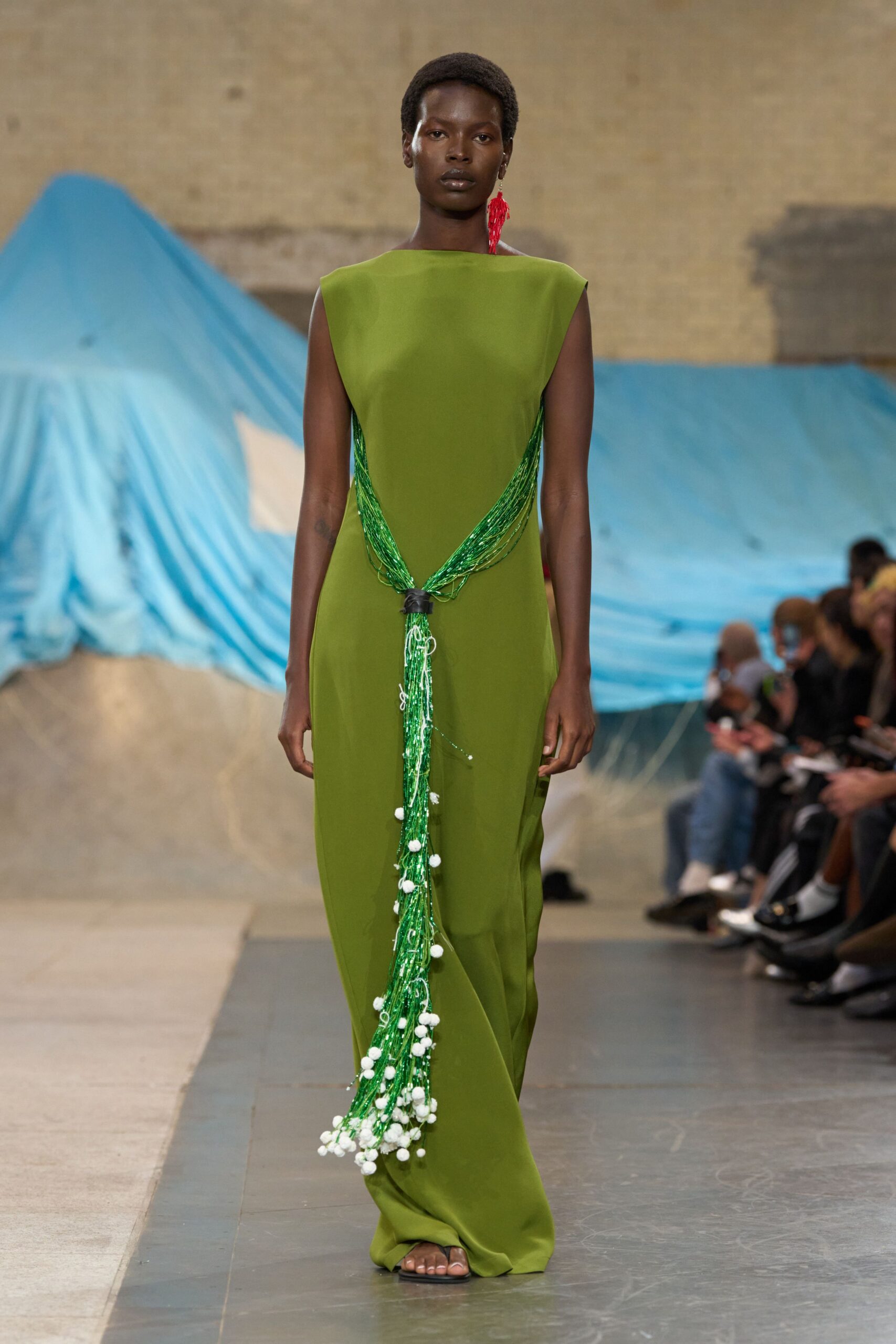 Olive column gown with a bateau neckline and beaded tassel cinching the waist. Single red tassel earring brushing the jaw. Flat black thong sandals barely peeking under the hem. Walking past tarped boats while the harbor smell clings to everything.