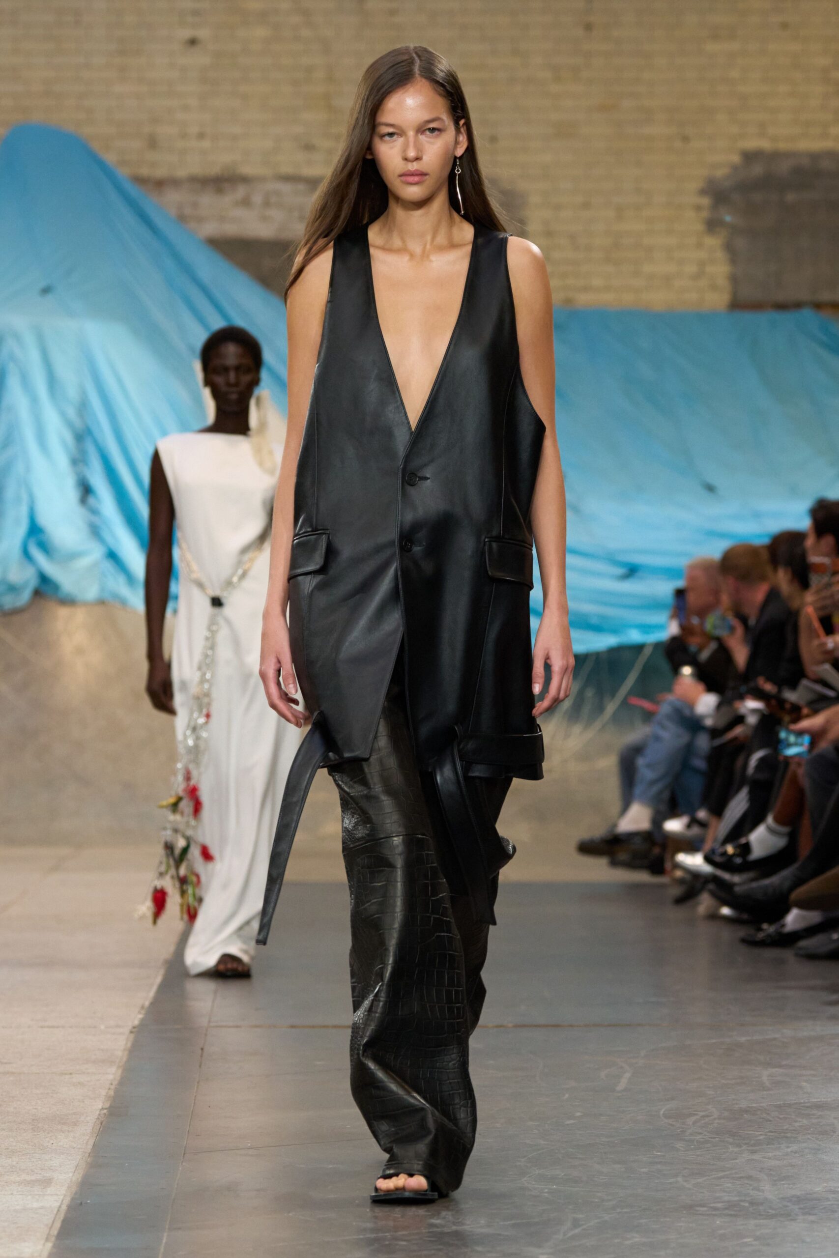 Single drop earring with a thin silver chain. Black leather long vest, plunging to the waist with one loose belt tab. Black crocodile-embossed leather trousers, cut wide and breaking over the foot. Square-toe black sandals. For crossing an empty convention hall before the booths open.